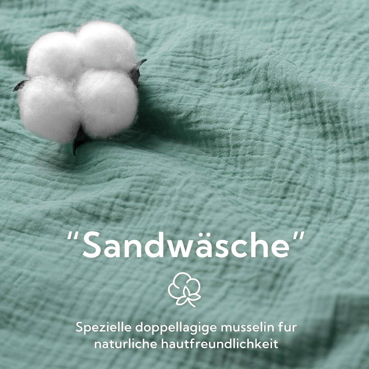 Ein Stück Baumwolle ruht auf einem strukturierten Stoffhintergrund. Text lautet: 'Sandwäsche'. Darunter ein Baumwoll-Icon mit Text: 'Spezielle doppellagige Musselin für natürliche Hautfreundlichkeit'.