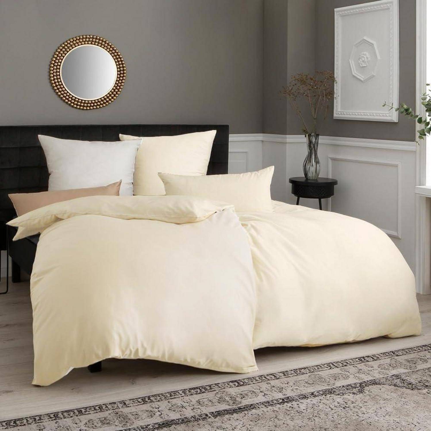 Ein Schlafzimmer mit einem ordentlich aufgemachten Bett, das mit cremefarbenen Leintüchern bezogen ist, mehreren Kissen, einem kleinen runden Spiegel an einer grauen Wand und einer minimalistischen Pflanzenvase.