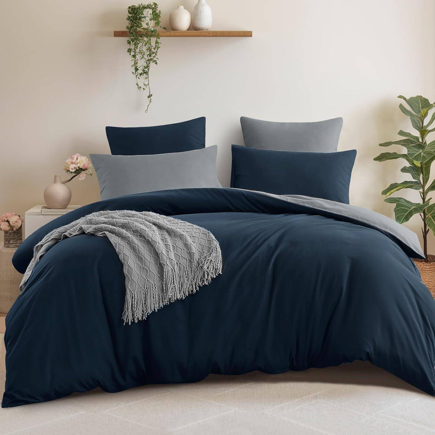 Ein ordentlich gemachtes Bett mit dunkelblauer Bettwäsche, zwei großen Kissen und einer grauen Decke. Eine kleine Pflanze und Dekoration befinden sich auf einem Regal darüber.
