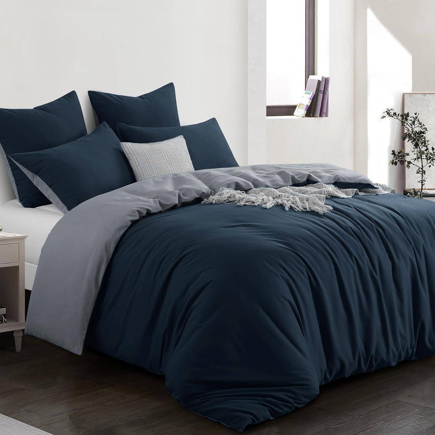 Ein sauber gemachtes Bett mit dunkelblauer Bettwäsche und mehreren Kissen ist in einem hellen Raum zu sehen, in dem rechts zwei Fenster Sonnenlicht hereinlassen.