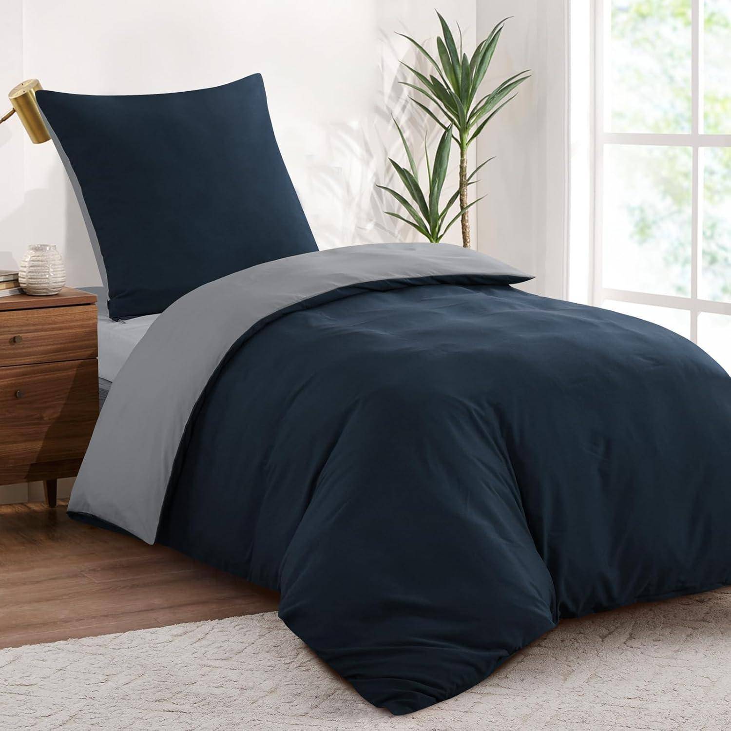 Ein sauber gemachtes Bett mit einer marineblau Bettdecke und Kissen, platziert in der Nähe eines Fensters. Ein kleiner Holznachttisch trägt eine Lampe und eine weiße Tasse.