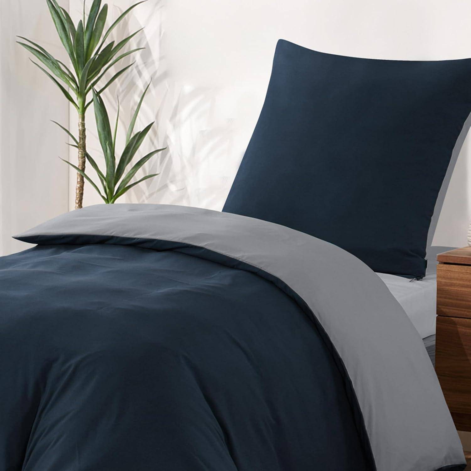 Ein sauber gemachtes Bett mit dunkelblauer Bettwäsche und passenden Kissen, neben einem hellholzfarbenen Nachttisch mit einer Topfpflanze daneben.
