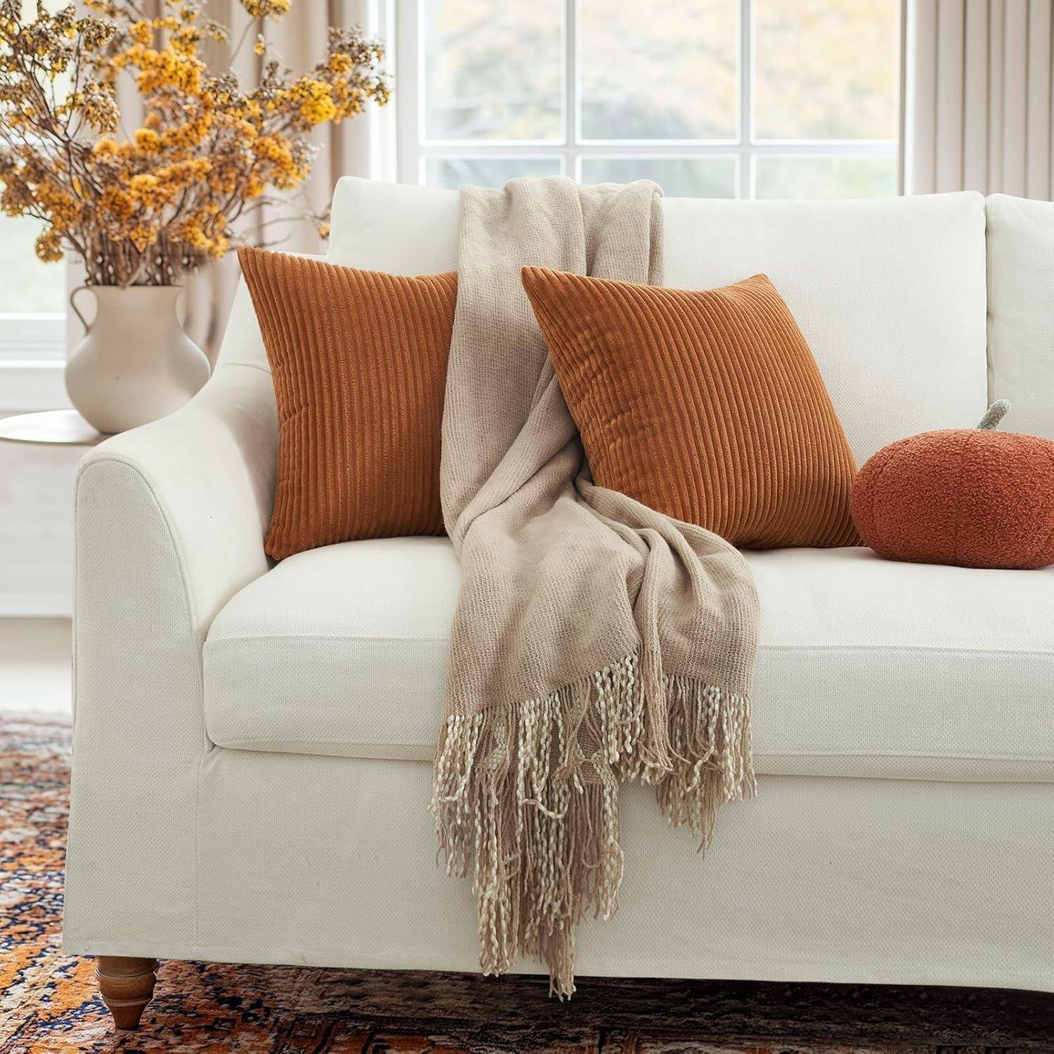 Ein weißes Sofa mit zwei braunen Kissen, einer beigen Decke und einem kleinen runden Kissen. Eine Vase mit herbstlichen Blumen steht auf einem Beistelltisch.