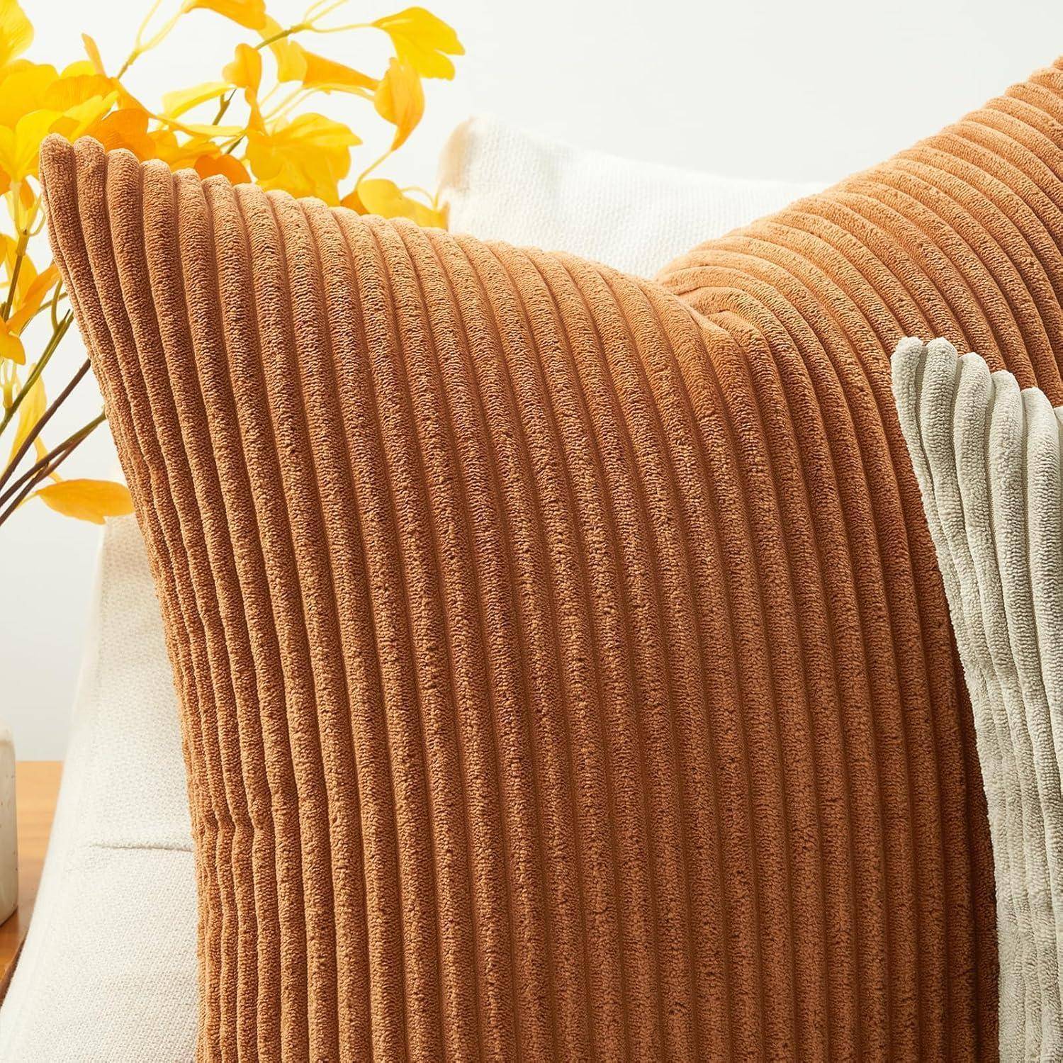Eine Nahaufnahme von zwei strukturierten Zierkissen, eines in Orange und eines in Grau, auf einem Sofa. Ein dekorativer Zweig mit gelben Blättern befindet sich im Hintergrund.