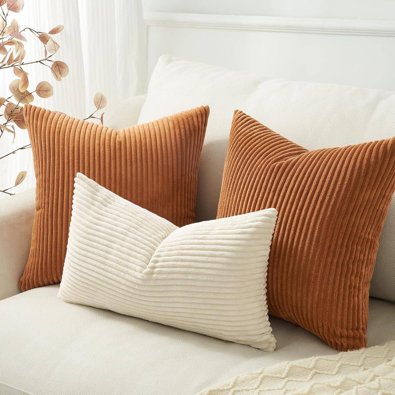 Drei Cordkissen in Creme- und Rostfarben sind auf einem weißen Sofa angeordnet und sind mit getrockneten Pflanzen in der Nähe dekoriert.