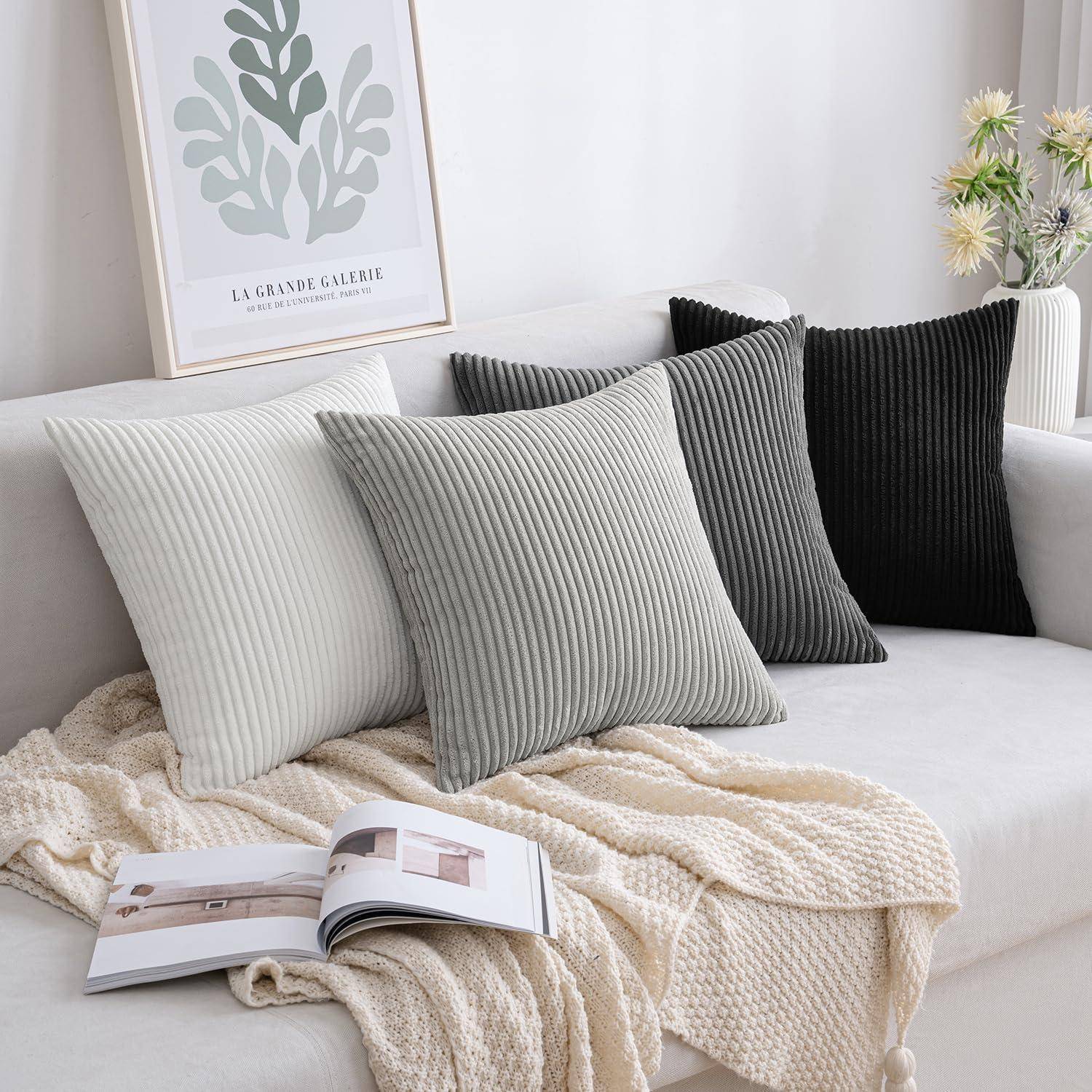 Eine gemütliche weiße Couch mit drei gerippten Kissen in Creme, Grau und Dunkelgrau, ein aufgeschlagenes Buch und ein Bild mit Pflanzenmuster darüber.