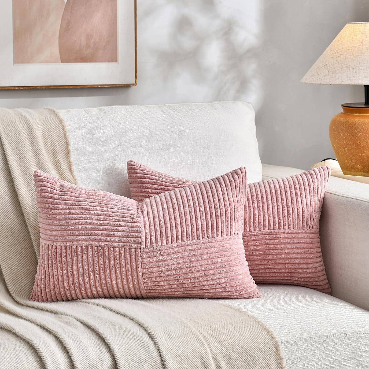 Zwei rosa strukturierte Kissen auf einem weißen Sofa mit einer beigen Decke. Eine Lampe und abstrakte Wandkunst befinden sich im Hintergrund.