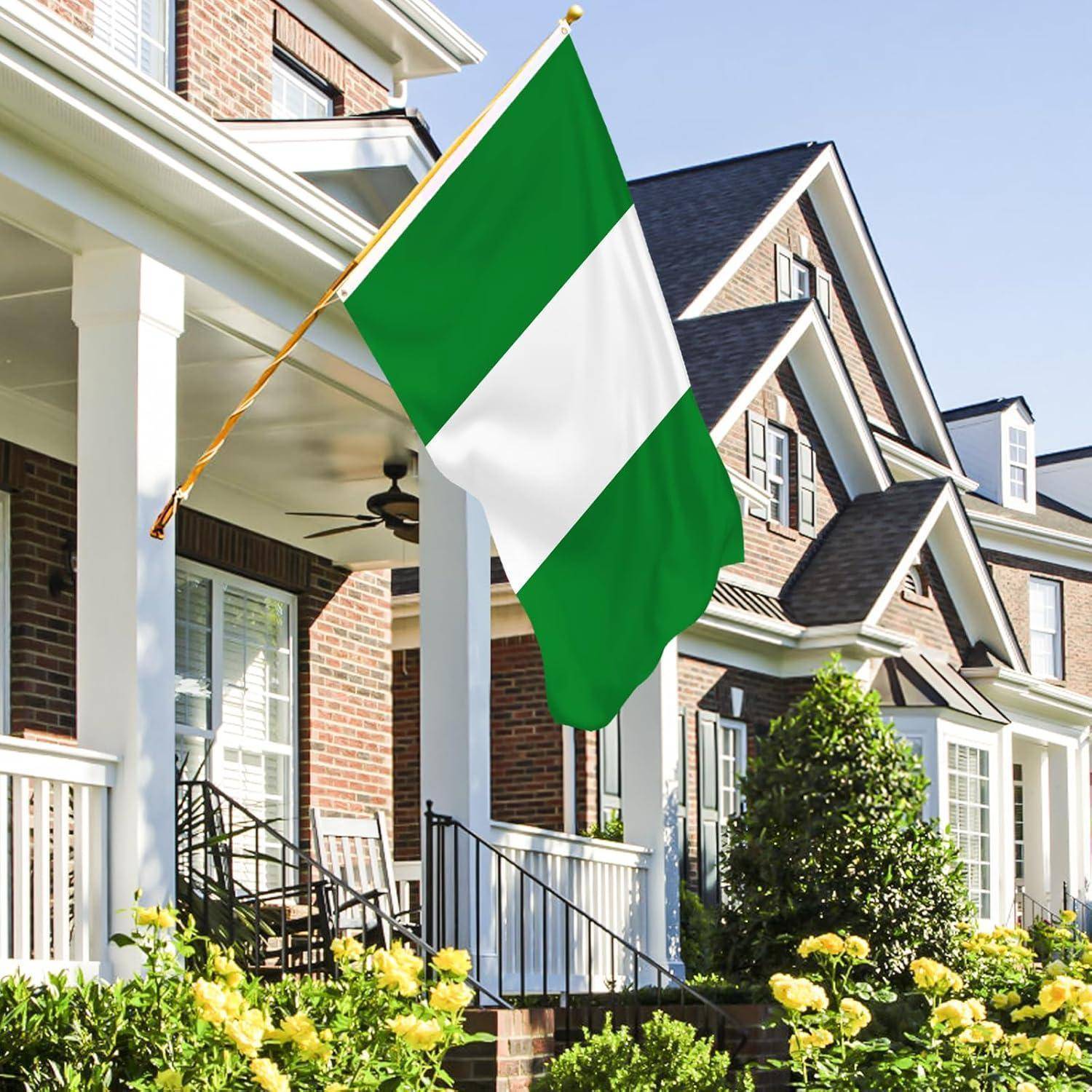 Nigerianische Flagge weht auf der Veranda eines Vorstadthauses, umgeben von gelben Blumen und Grünpflanzen, vor einer Kulisse ähnlicher Häuser.