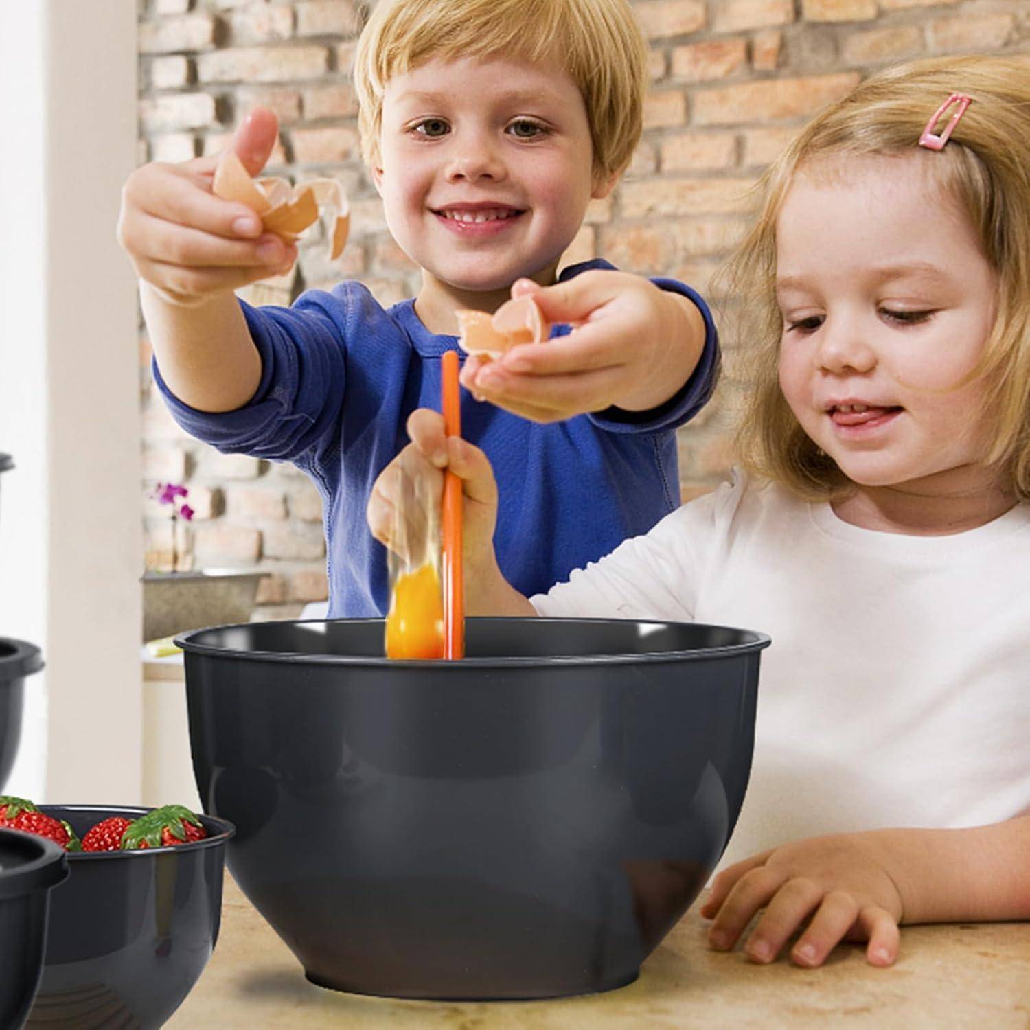 Zwei Kinder lächeln, während sie Eier in eine Schüssel schlagen, in einer Küche. Eine Schüssel mit Erdbeeren ist im Vordergrund sichtbar.