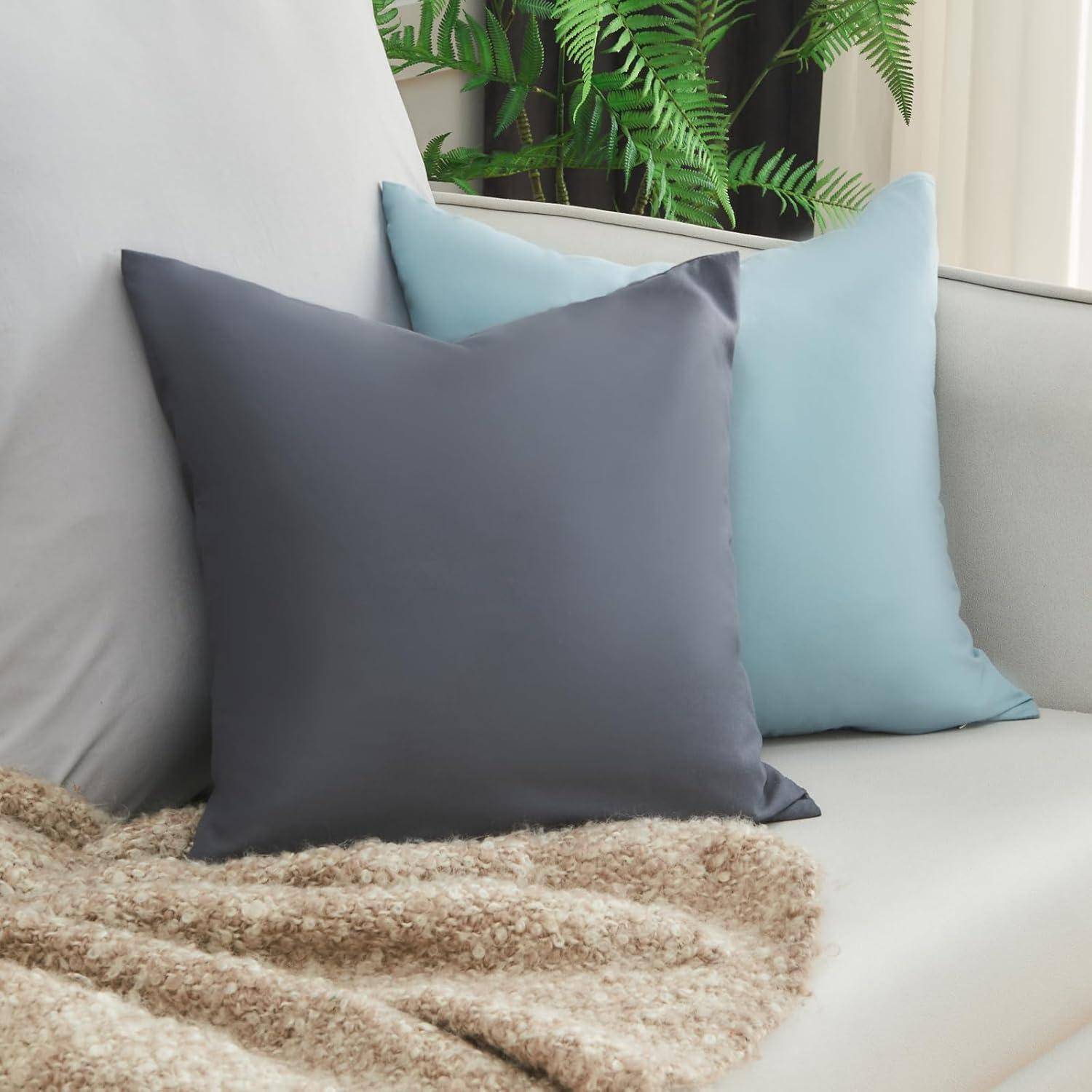 Eine gemütliche Sofa-Szene mit zwei großen Kissen in Grau- und Blautönen und einer flauschigen beigen Decke. Eine grüne Pflanze im Hintergrund verleiht der Szene eine Note von Natürlichkeit.