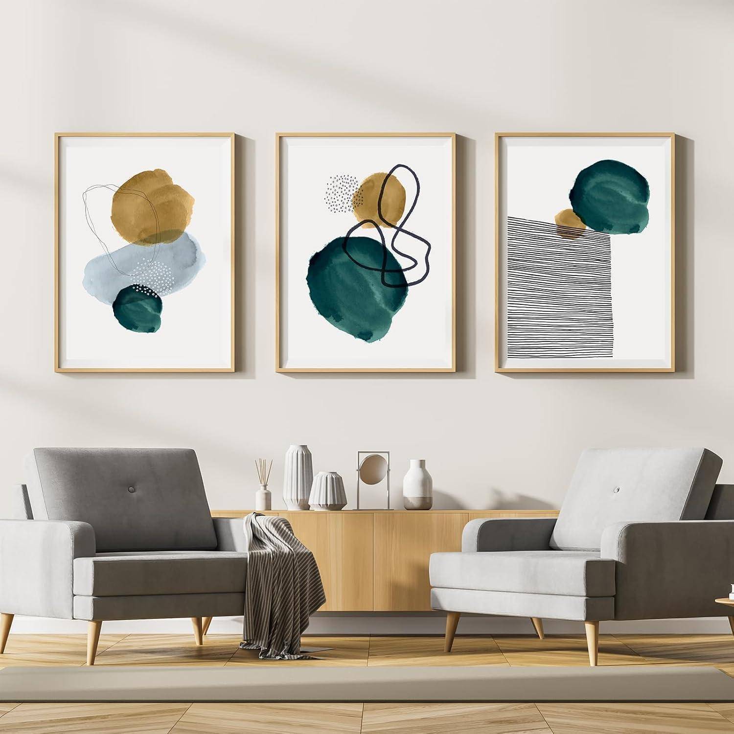 Ein moderner Wohnraum mit zwei grauen Sesseln und einem Holzsideboard unter drei abstrakten Kunstwerken in Grün, Gold und Schwarz.
