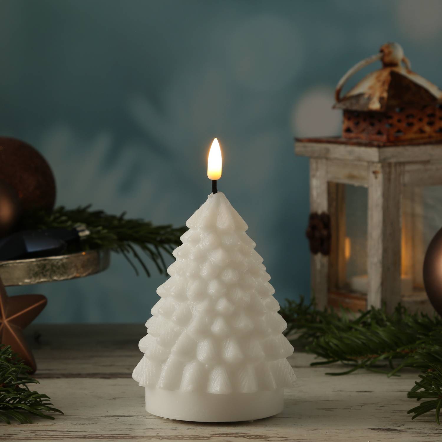 Eine brennende Kerze in Form eines Tannenbaums steht auf einer Holzoberfläche, umgeben von Immergrünzweigen, mit einer Laterne im Hintergrund.