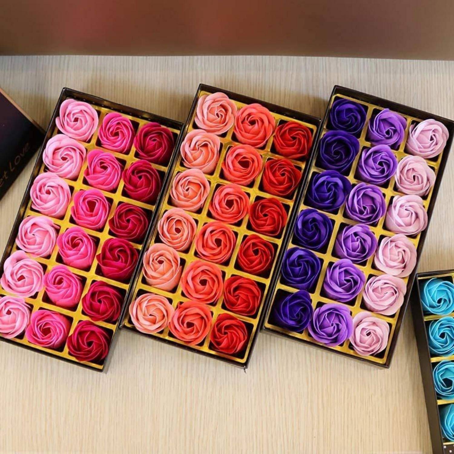 Vier rechteckige Kartons, gefüllt mit rosenförmiger Seife in verschiedenen Schattierungen von Rosa, Rot und Lila, sorgfältig auf einer Holzoberfläche angeordnet.