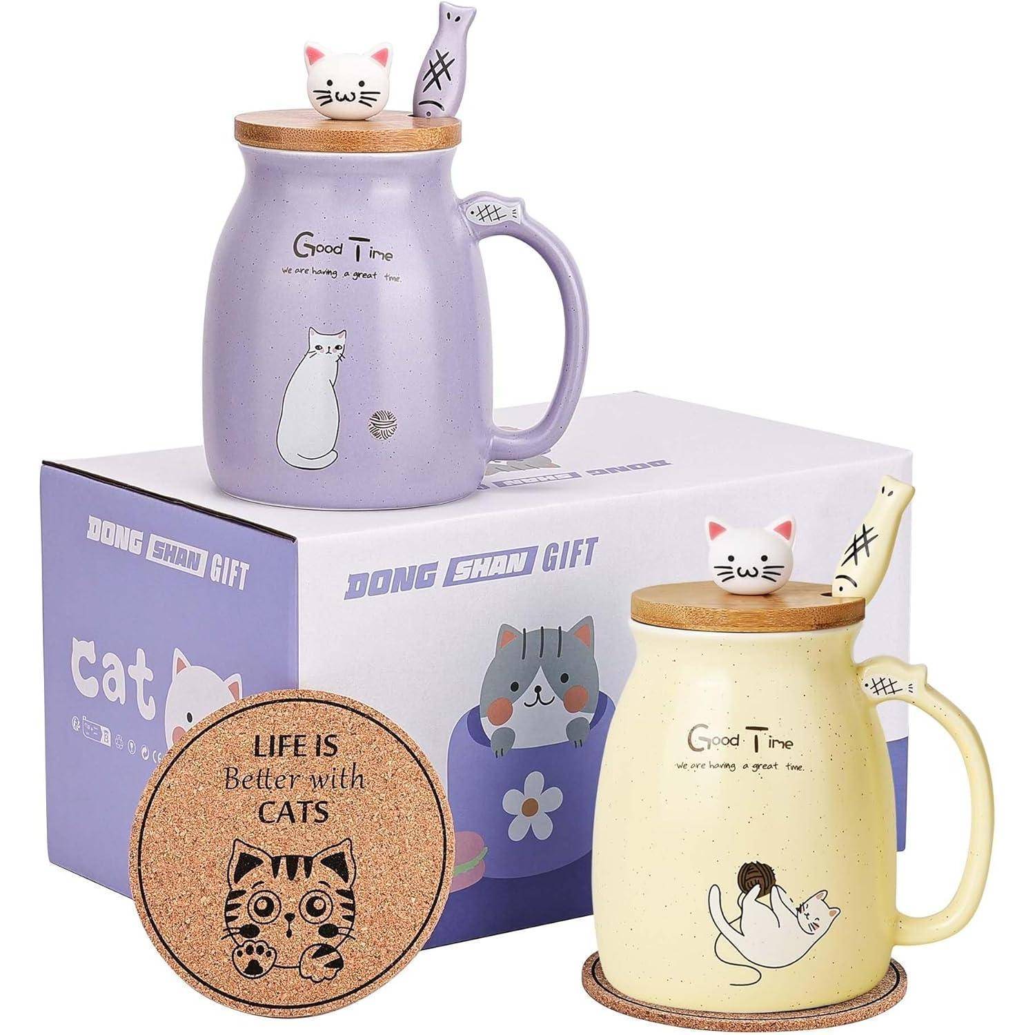 Zwei Katzen-Tassen, eine in Lila und eine in Cremeweiß, mit passenden Deckeln und Löffeln, neben einem Untersetzer aus Kork mit der Aufschrift ‚Das Leben ist besser mit Katzen'.