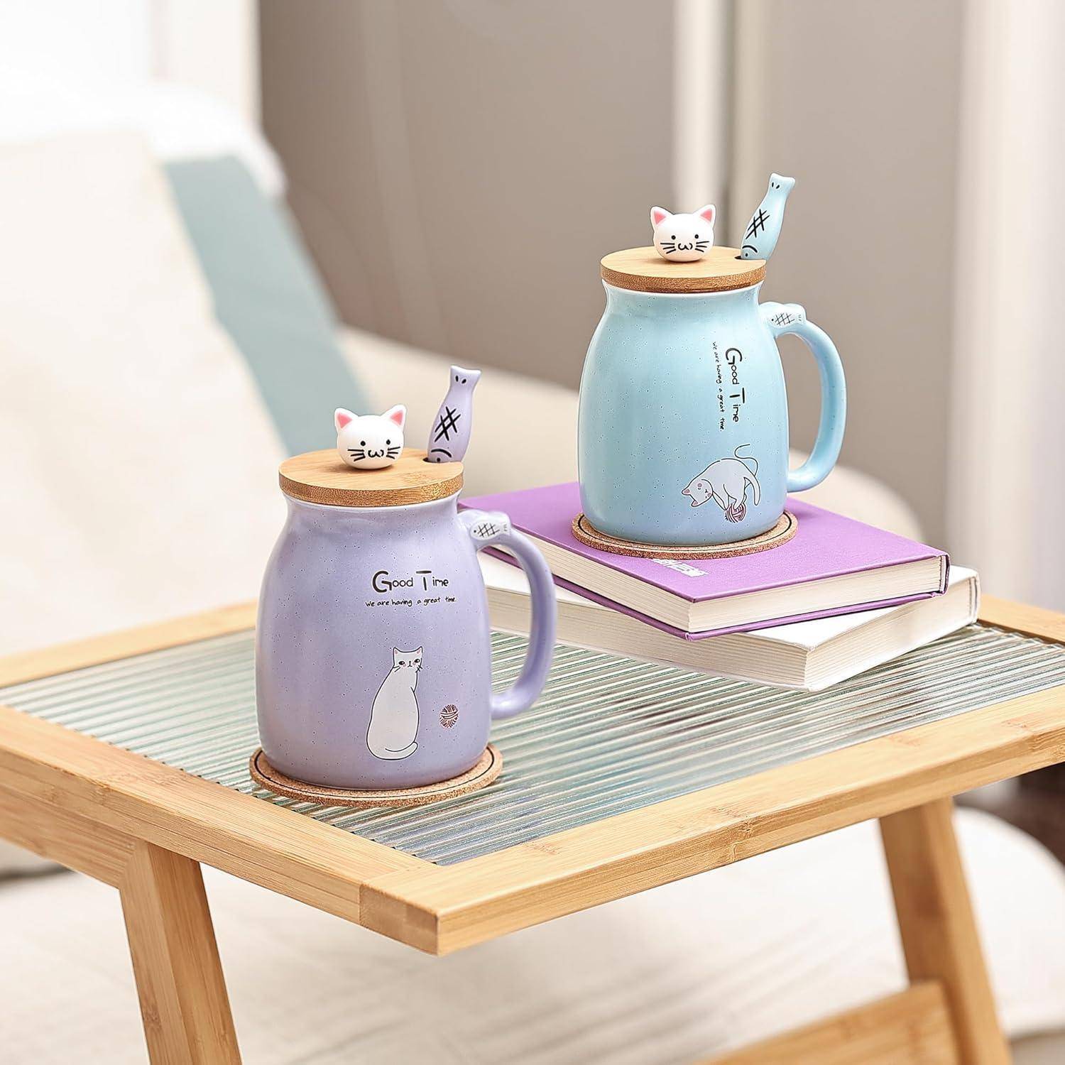 Zwei Keramiktassen mit Katzendesigns und Deckeln stehen auf einem Holztablett. Eine blaue Tasse steht auf einem Stapel Bücher, und eine lila Tasse steht auf Untersettern.