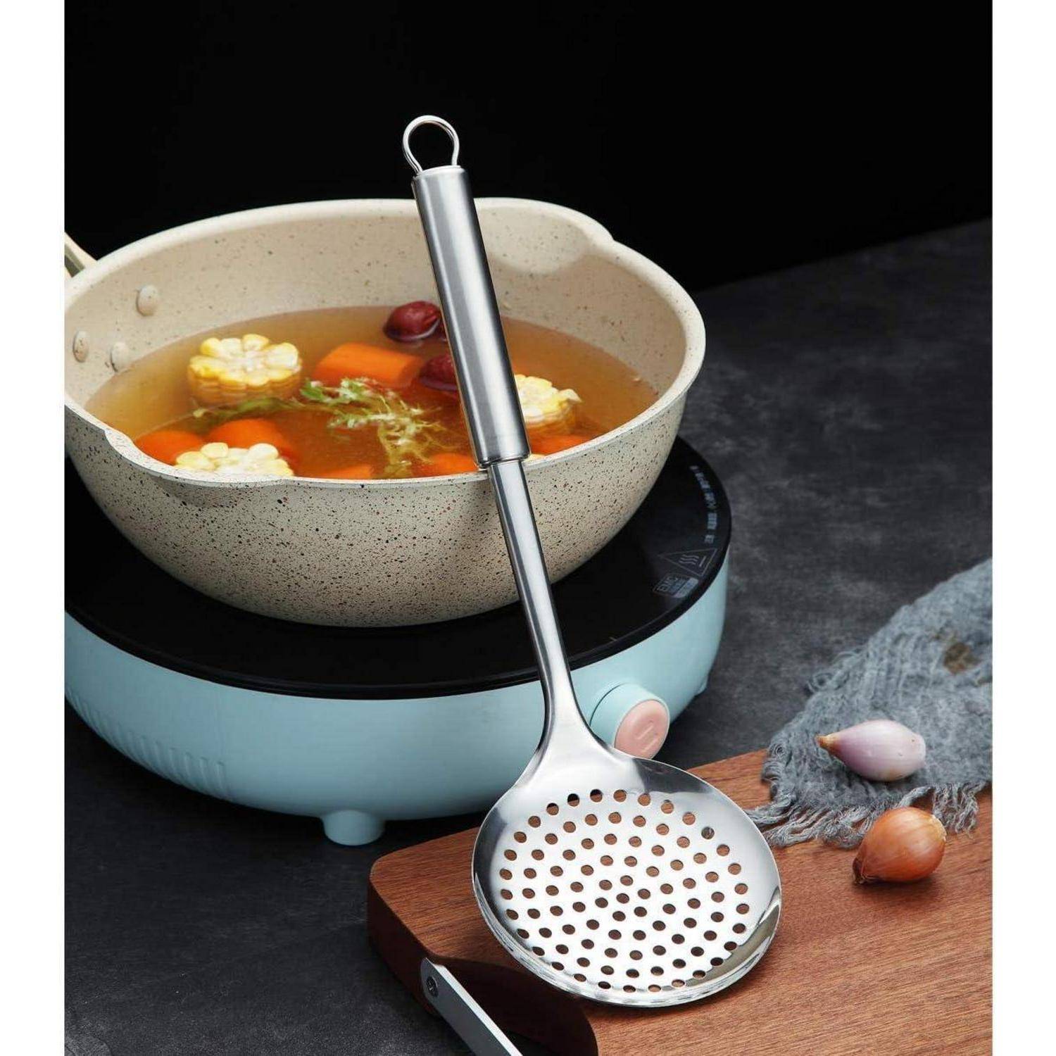 Ein Metalllöffel zum Abschöpfen liegt auf einer dunklen Arbeitsplatte neben einem Topf mit Gemüsesuppe, der Mais, Karotten und Grüngemüse enthält, auf einem Herd.