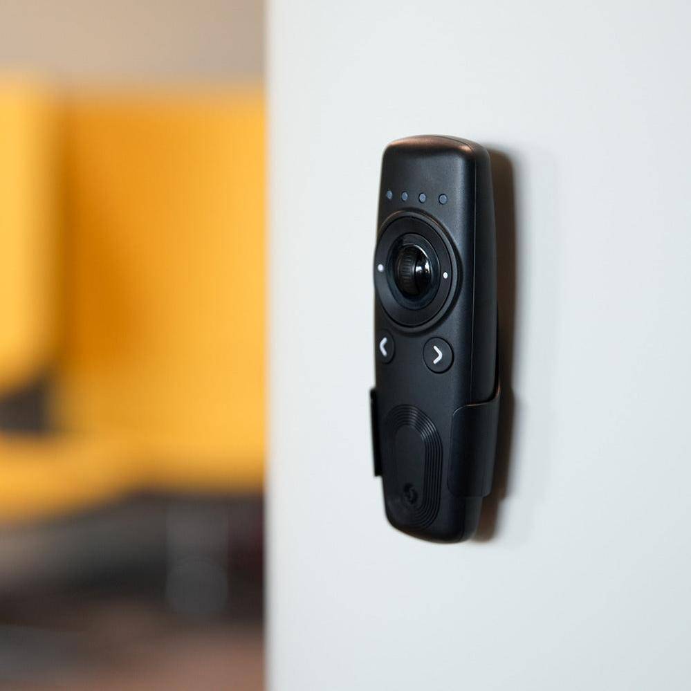 Ein schwarzes elektronisches Gerät mit Tasten und Kameraobjektiv ist an einer weißen Wand montiert. Ein unscharfer gelber Stuhl befindet sich im Hintergrund.