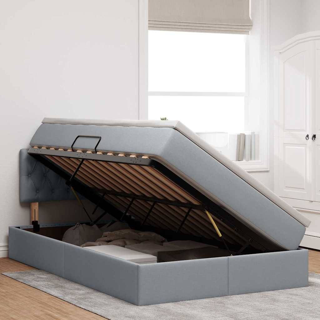 Ein schlankes, modernes Bett mit grauem Polster, bei dem die Matratze angehoben ist, um Stauraum darunter zu offenbaren, in einem hellen Raum mit weißen Wänden und einem Fenster.