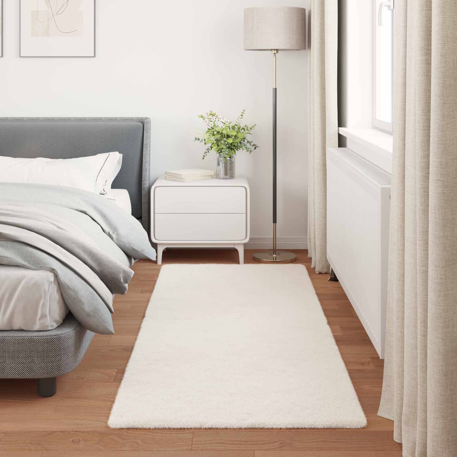 Ein modernes Schlafzimmer mit einem grauen Bett, einem weißen Nachttisch, einer Lampe und einer Pflanze. Helle Vorhänge und ein weißer Teppich sorgen für eine lichte, luftige Atmosphäre.