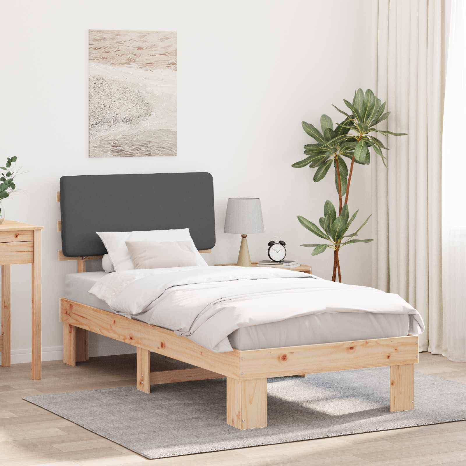 Ein minimalistisches Schlafzimmer mit einem Kiefernbett, grauem Kopfteil und weißer Bettwäsche. Ein Holznachttisch, eine Pflanze und eine Lampe ergänzen die schlichte Dekoration.