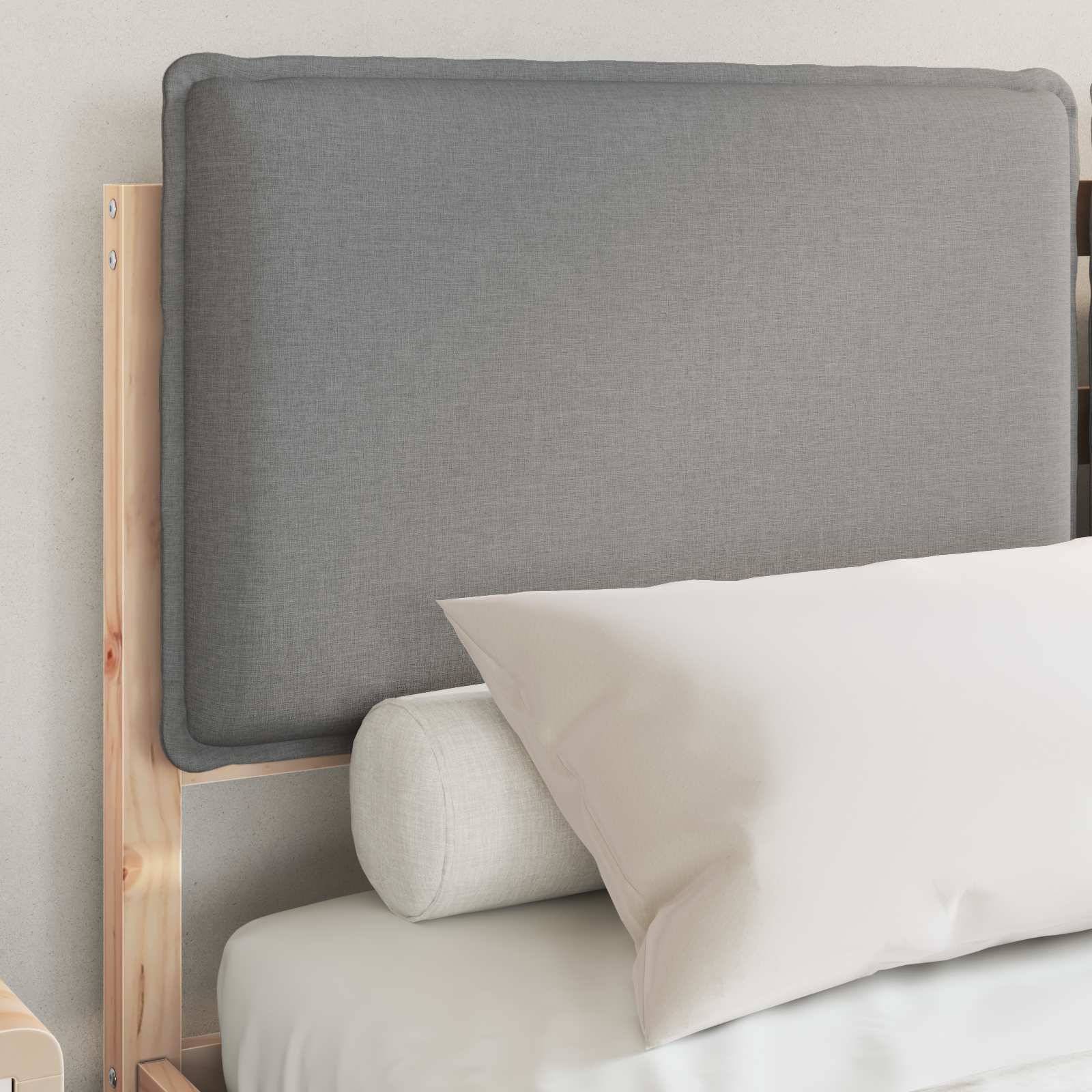 Ein ordentlich gemachtes Bett mit einem einfachen Holzrahmen, grauem Stoffkopfteil, weißem Kissen und einem zylindrischen Nackenrolle vor einer hellgefärbten Wand.