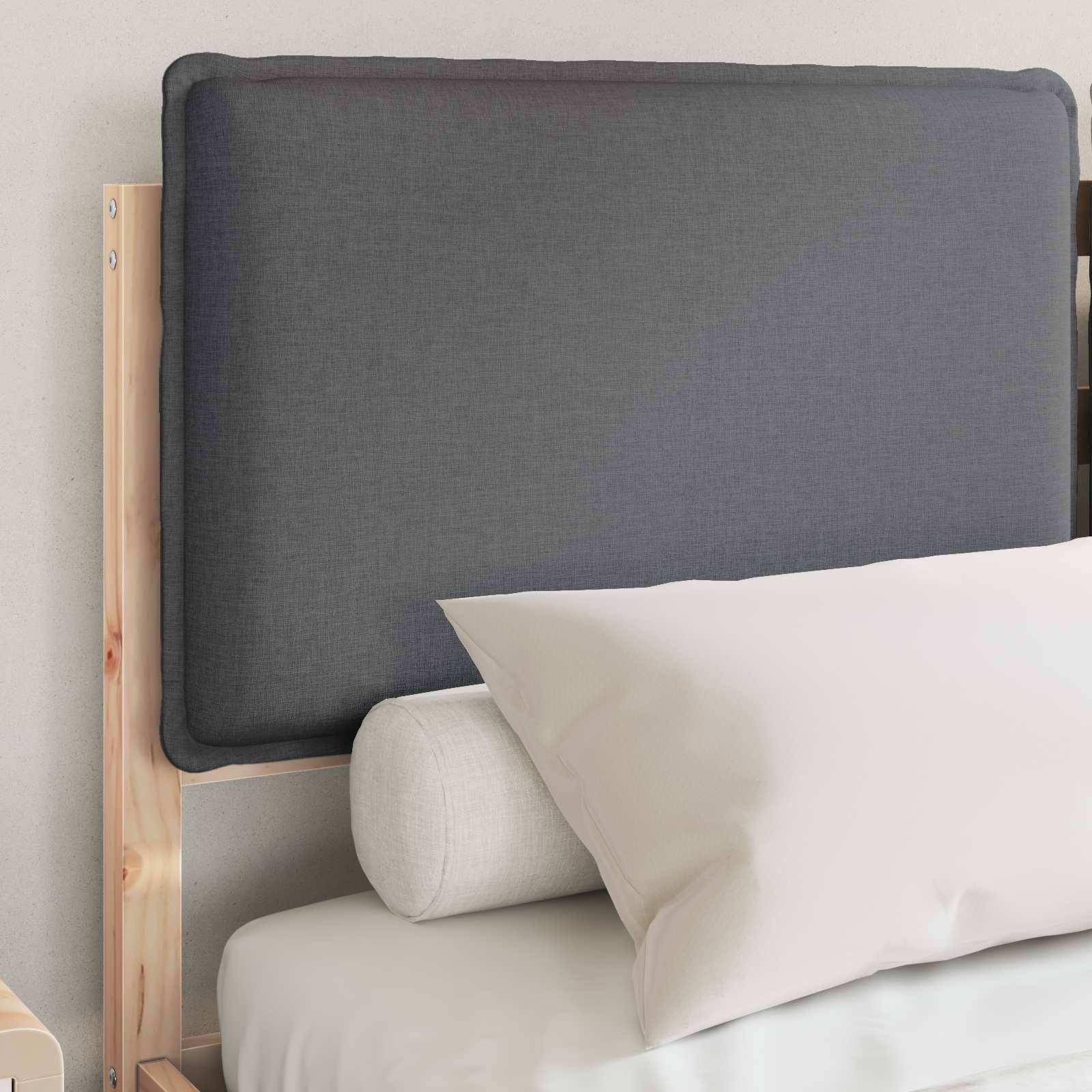 Ein Holzbettrahmen mit einem grau gepolsterten Kopfteil, einem großen weißen Kissen und einem kleineren zylindrischen Bolsterkissen vor einer hellen Wand.