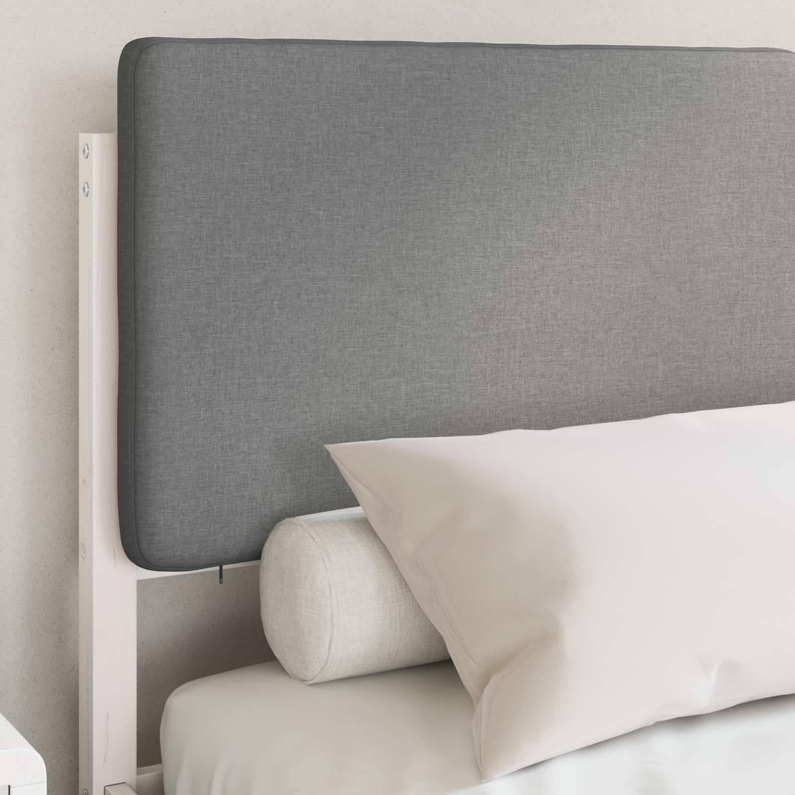 Graue gepolsterte Kopfende mit einem hellgefärbten Betthauptkissen und einem Nackenrolle auf einem weißen Bett, vor einer neutralen Wand.