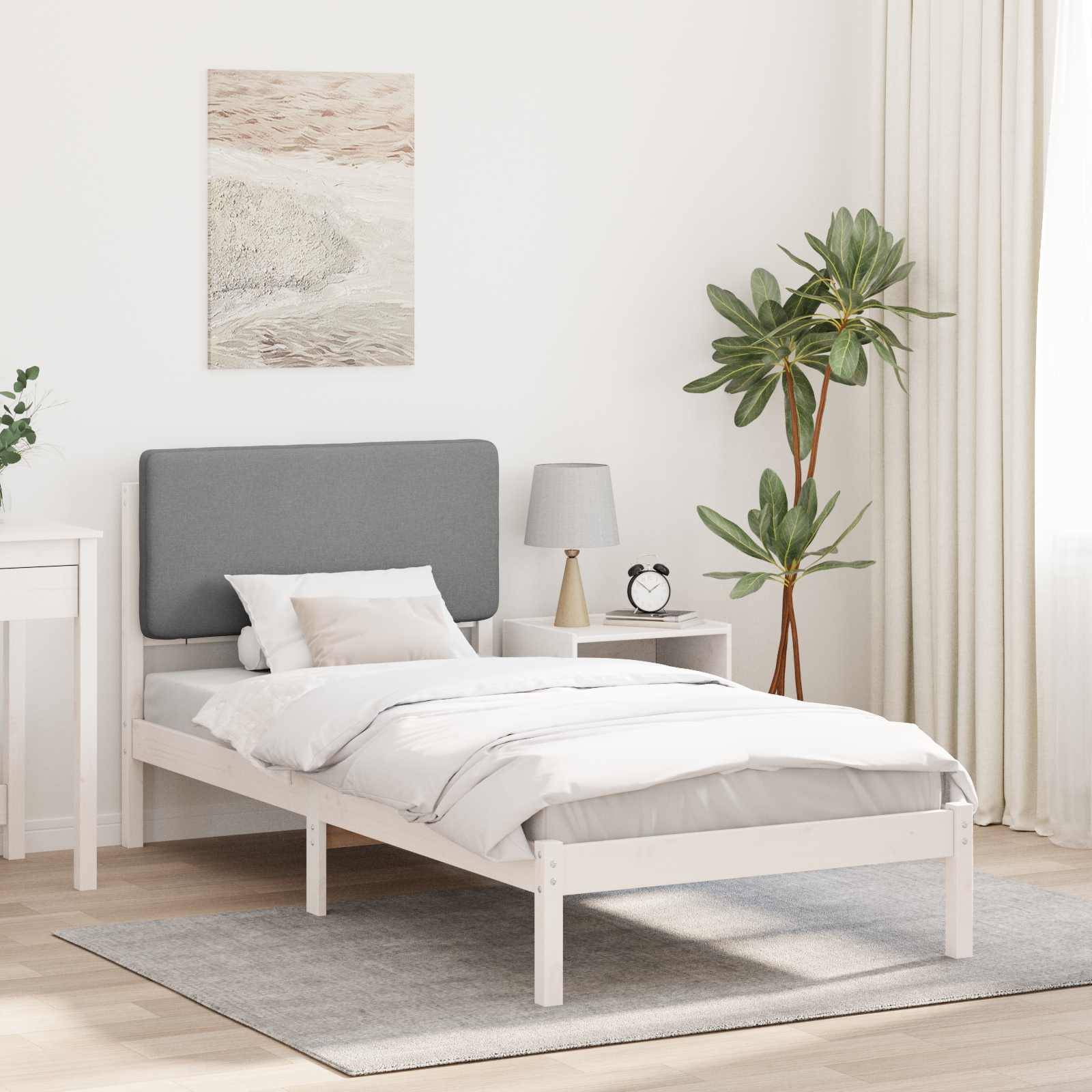 Ein minimalistisches Schlafzimmer mit einem weißen Bett, einem grauen Kopfteil und weißer Bettwäsche. Ein Nachttisch trägt eine Lampe und eine Pflanze. Eine blattreiche Pflanze steht neben einem Fenster mit transparenten Gardinen.