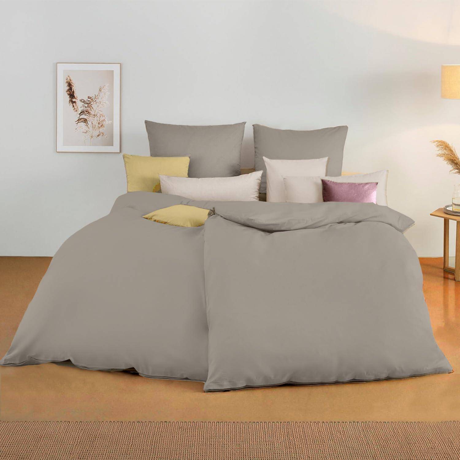 Ein Bett mit grauer Bettwäsche, mehreren Kissen in Beige- und Rosatönen, einem gerahmten Bild an der Wand und einer Lampe auf einem Beistelltisch.