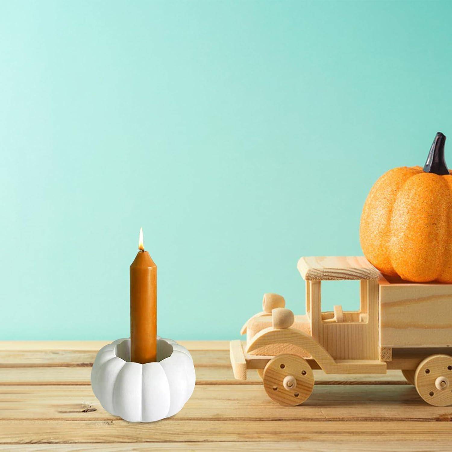 Ein kleiner Holzlastwagen mit einem Kürbis auf der Ladefläche steht auf einer Holzoberfläche. Links daneben befindet sich ein weißer, kürbisförmiger Kerzenhalter mit einer brennenden Kerze.