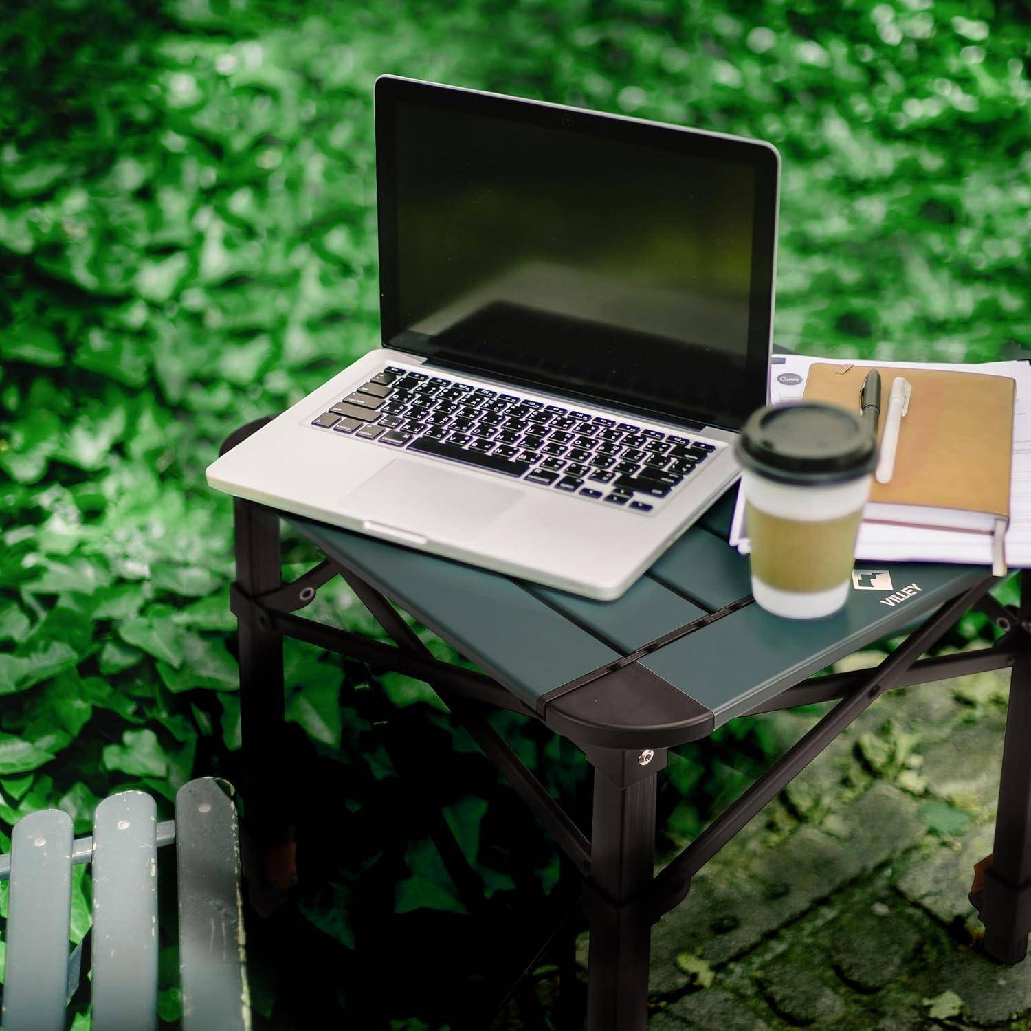 Ein Laptop auf einem kleinen Outdoor-Tisch neben einer Kaffeetasse und einem Notizbuch, mit belaubtem Hintergrund, was Arbeit oder Studium in einer Naturumgebung andeutet.