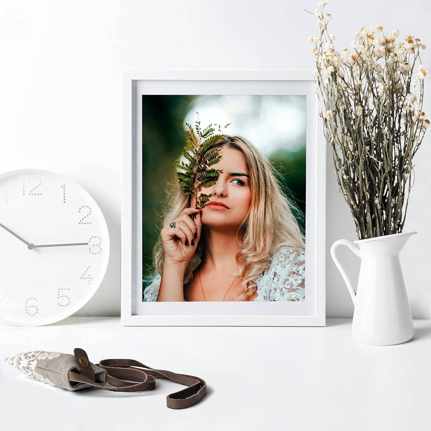 Ein gerahmtes Foto einer Frau, die ein Farnblatt hält, auf einem weißen Tisch neben einer Uhr, einer Vase mit getrockneten Blumen und einem kleinen Beutel.