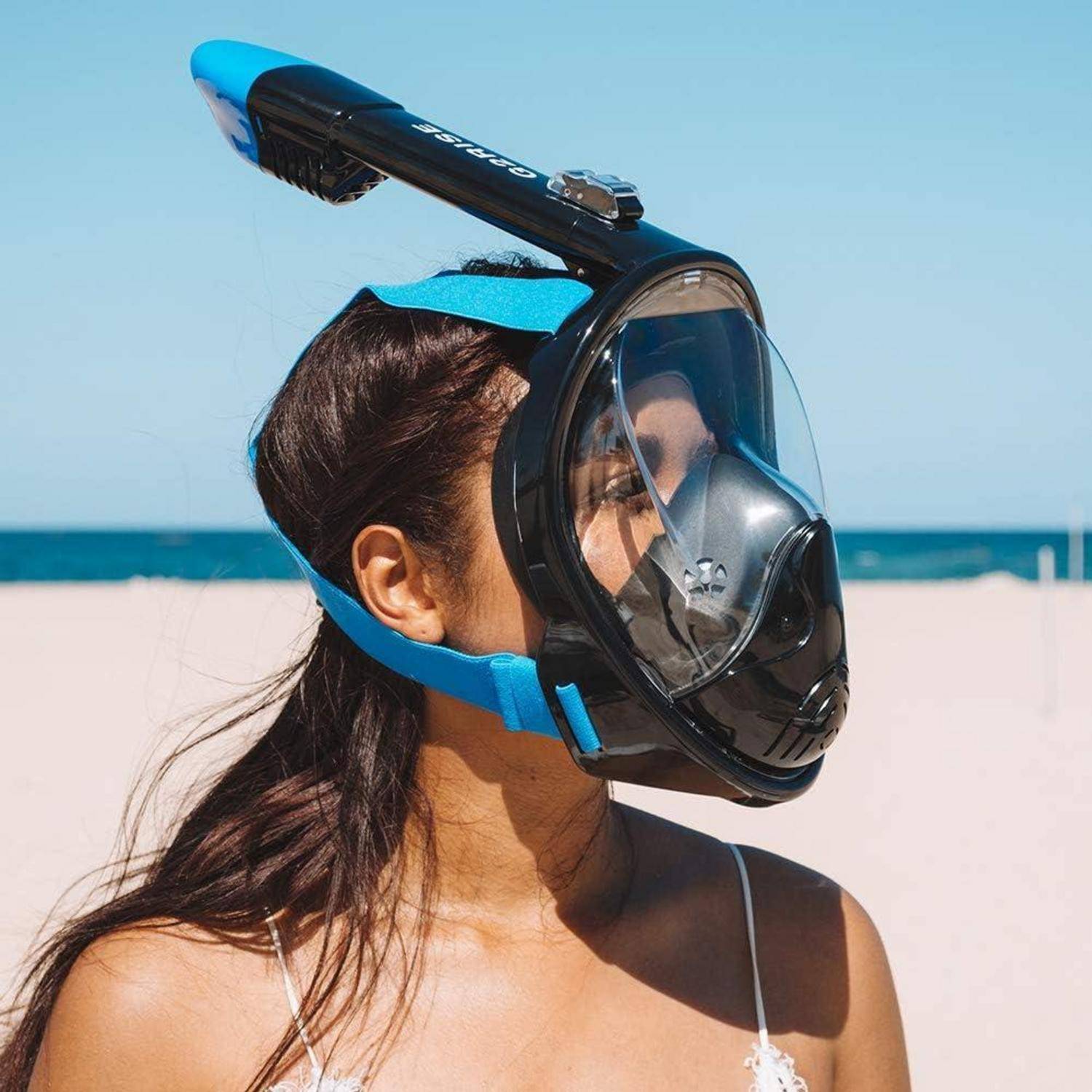 Eine Person trägt auf einem Strand eine Tauchmaske mit Vollgesichtsschutz und blickt zum Ozean. Der Himmel ist klar, und der Sand strahlt hell im Sonnenlicht.