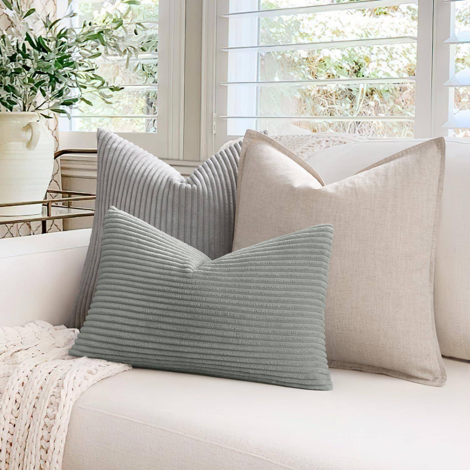 Ein gemütliches weißes Sofa mit zwei dekorativen Kissen, einem grauen gerippten und einem beigen, neben einer weichen gestrickten Decke, vor einer Fensterkulisse.