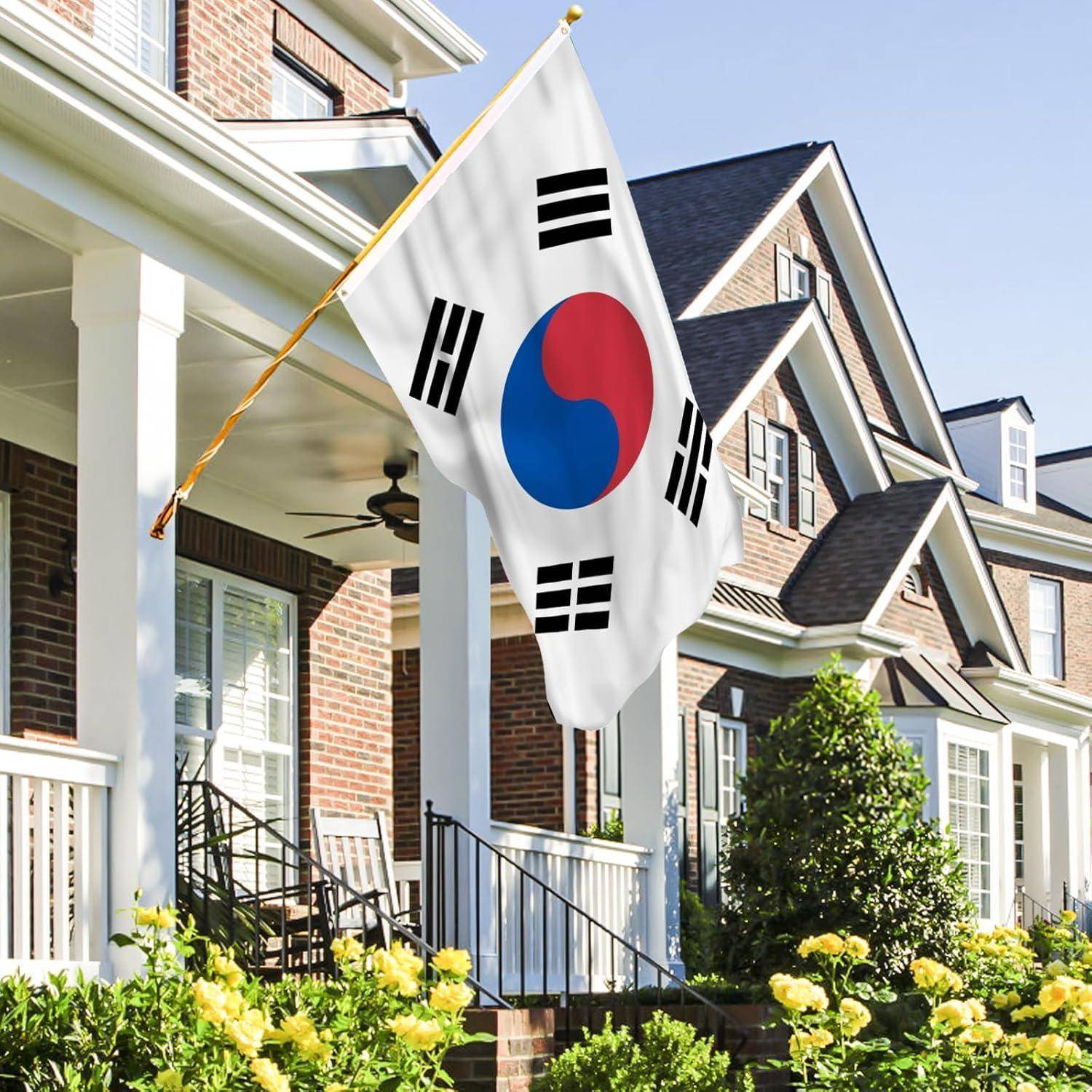 Eine südkoreanische Flagge hängt vor einem Backsteinhaus in einem Vorort, mit einer Veranda und blühenden gelben Blumen im Vordergrund.