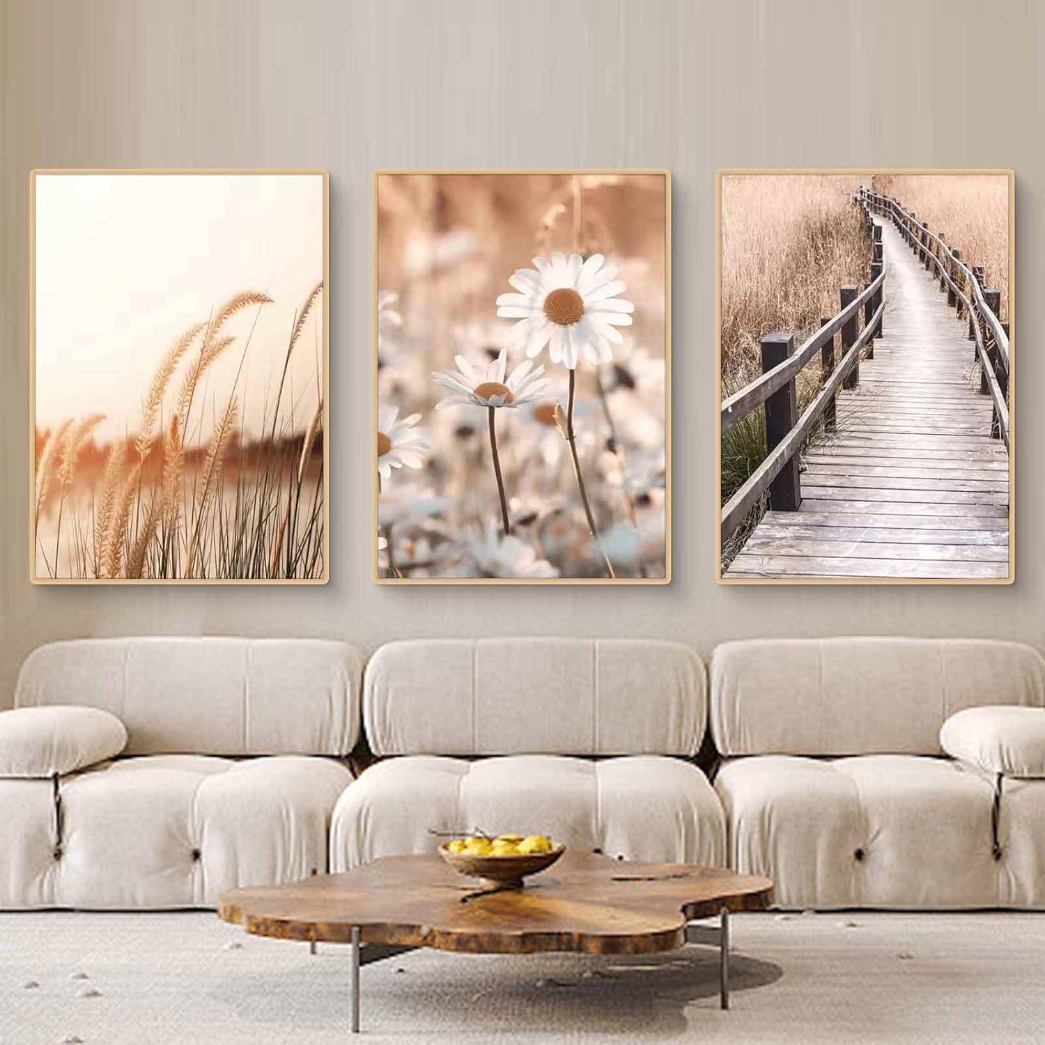 Drei gerahmte Naturfotos über einem beigen Sofa: Gräser bei Sonnenaufgang, weiße Gänseblümchen und ein hölzerner Steg durch hohe Schilfrohrbestände.