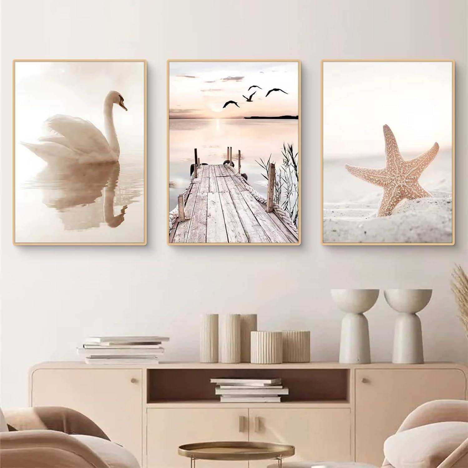Drei gerahmte Kunstdrucke: ein Schwan im Wasser, ein Steg bei Sonnenuntergang mit Vögeln und ein Seestern auf Sand, schmücken eine beige Wand über Regalen.
