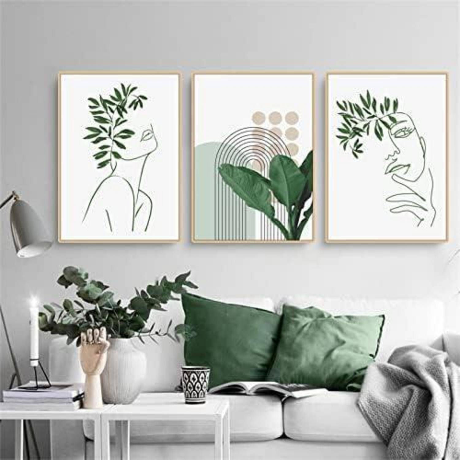 Drei minimalistische Linienkunst-Drucke von Gesichtern und Pflanzen an der Wand über einem weißen Sofa mit grünen Kissen, neben einer Lampe und Topfpflanzen.