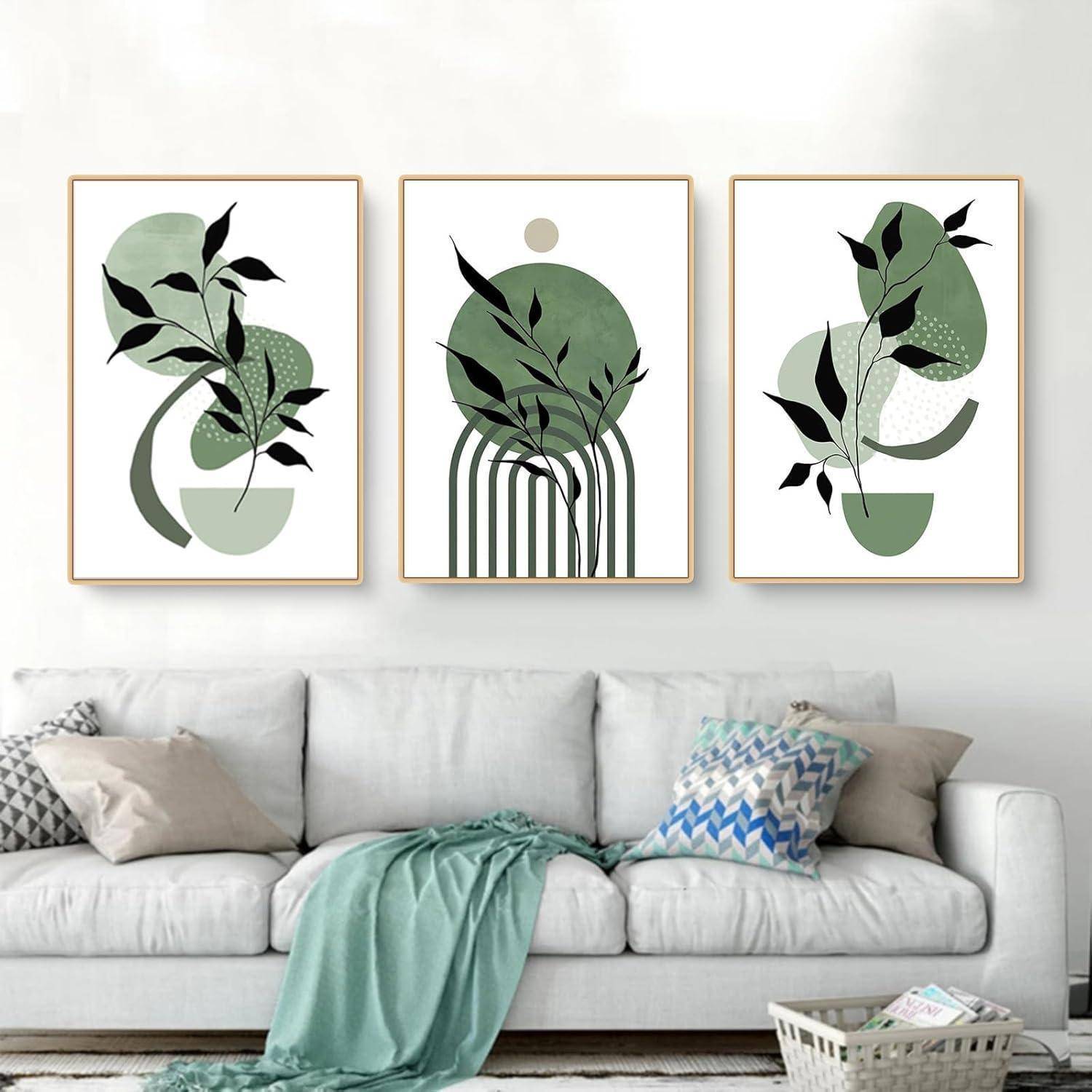 Ein moderner Wohnraum mit einem weißen Sofa und einer grünen Decke. Darüber befinden sich drei minimalistische abstrakte Kunstwerke mit grünen Schattierungen und schwarzen Blattmotiven.