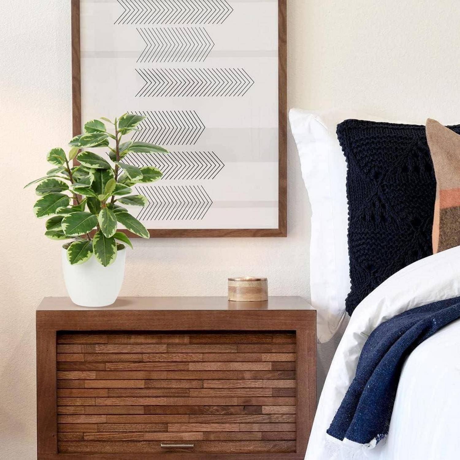 Ein hölzerner Nachttisch mit einer Topfpflanze und gerahmter geometrischer Kunstarbeit neben einem Bett mit einem marineblauem Kissen und weißen Laken.