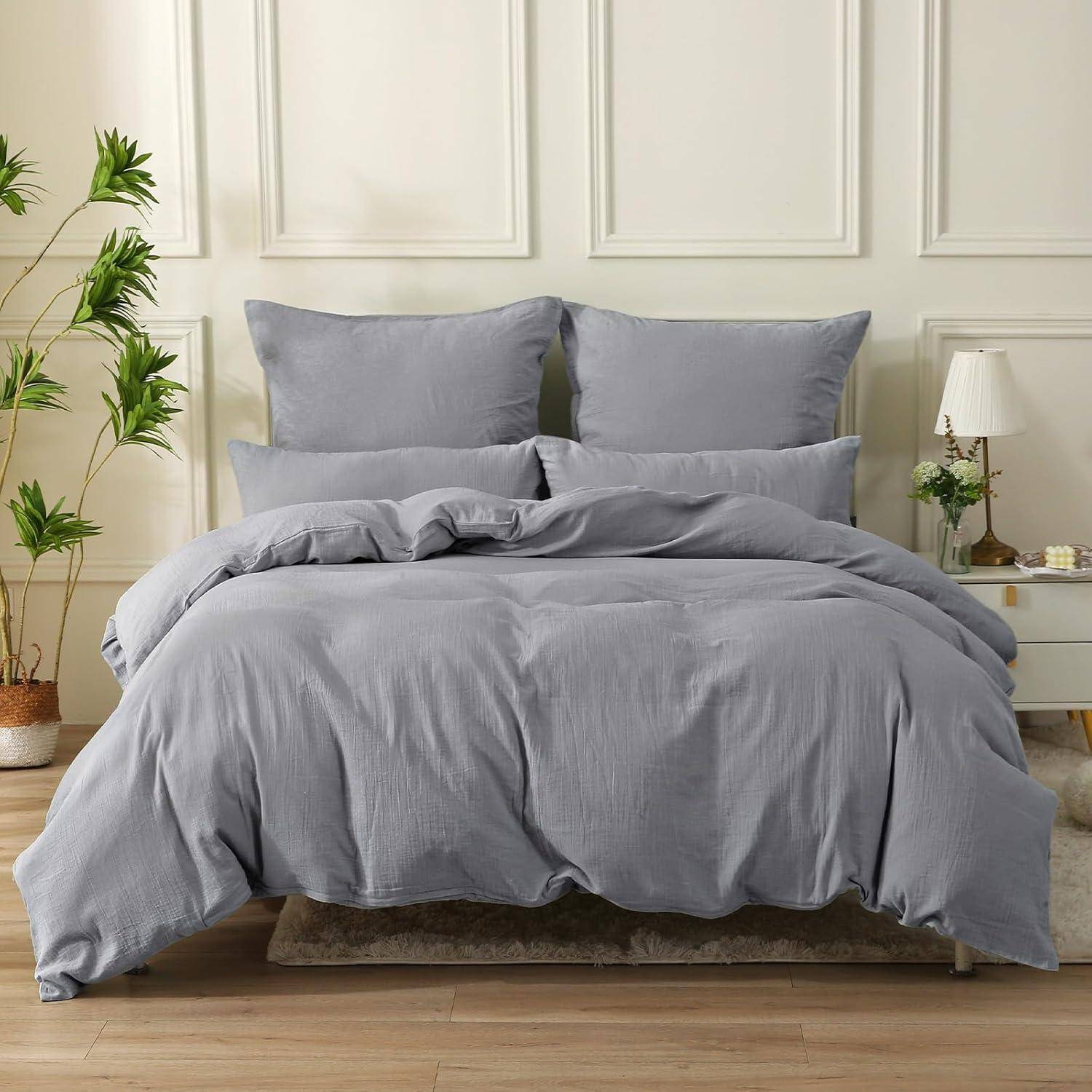 Ein graues Bettwäscheset mit Kissen und Bettdecke auf einem Bett, neben einem Nachttisch und einer Pflanze, in einem hellgefärbten, eleganten Schlafzimmer.