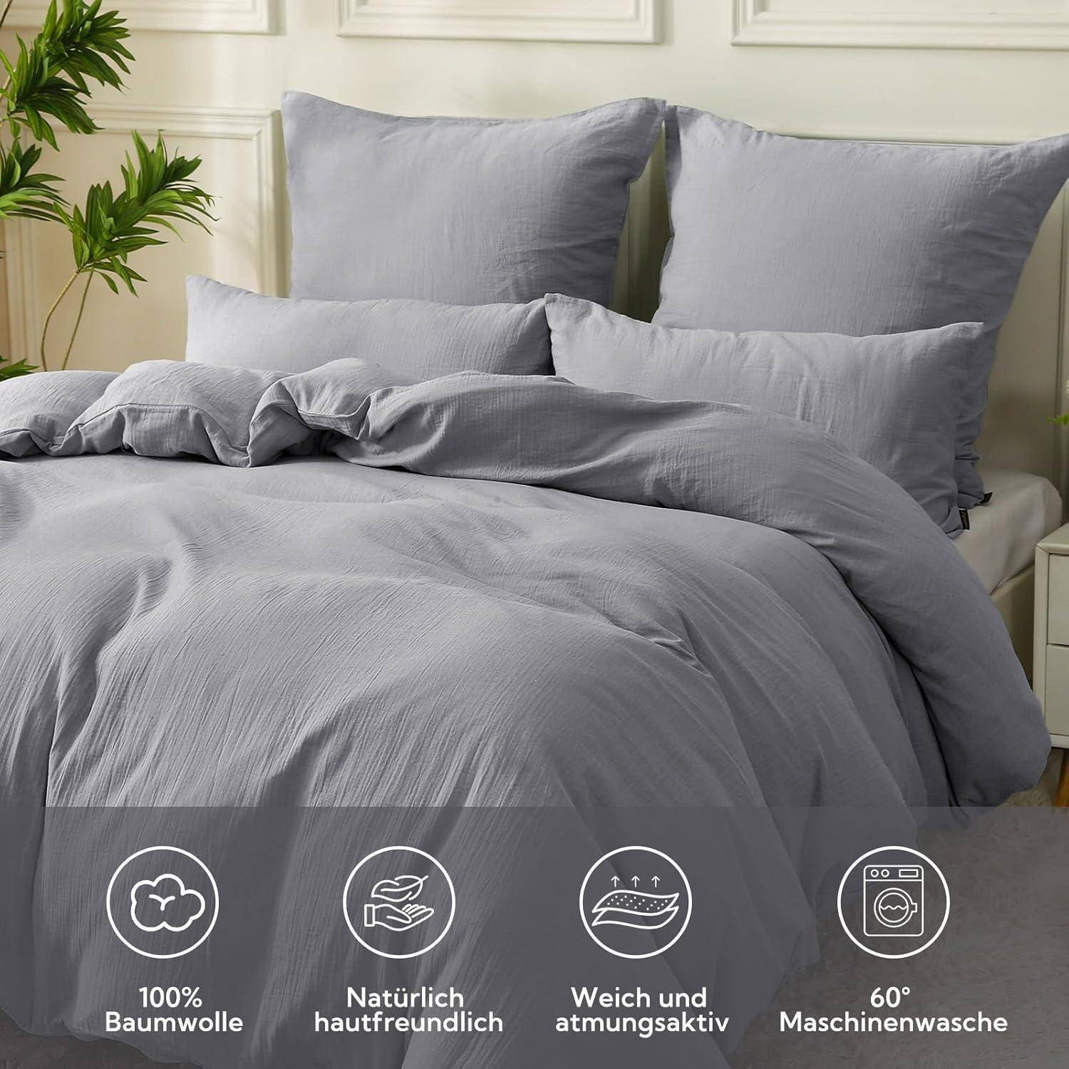 Ein sauber gemachtes Bett mit grauer Bettwäsche, bestehend aus Bettdecke, Kissen und Laken. Icons darunter zeigen Eigenschaften: 100% Baumwolle, hautfreundlich, atmungsaktiv, bei 60°C waschbar.