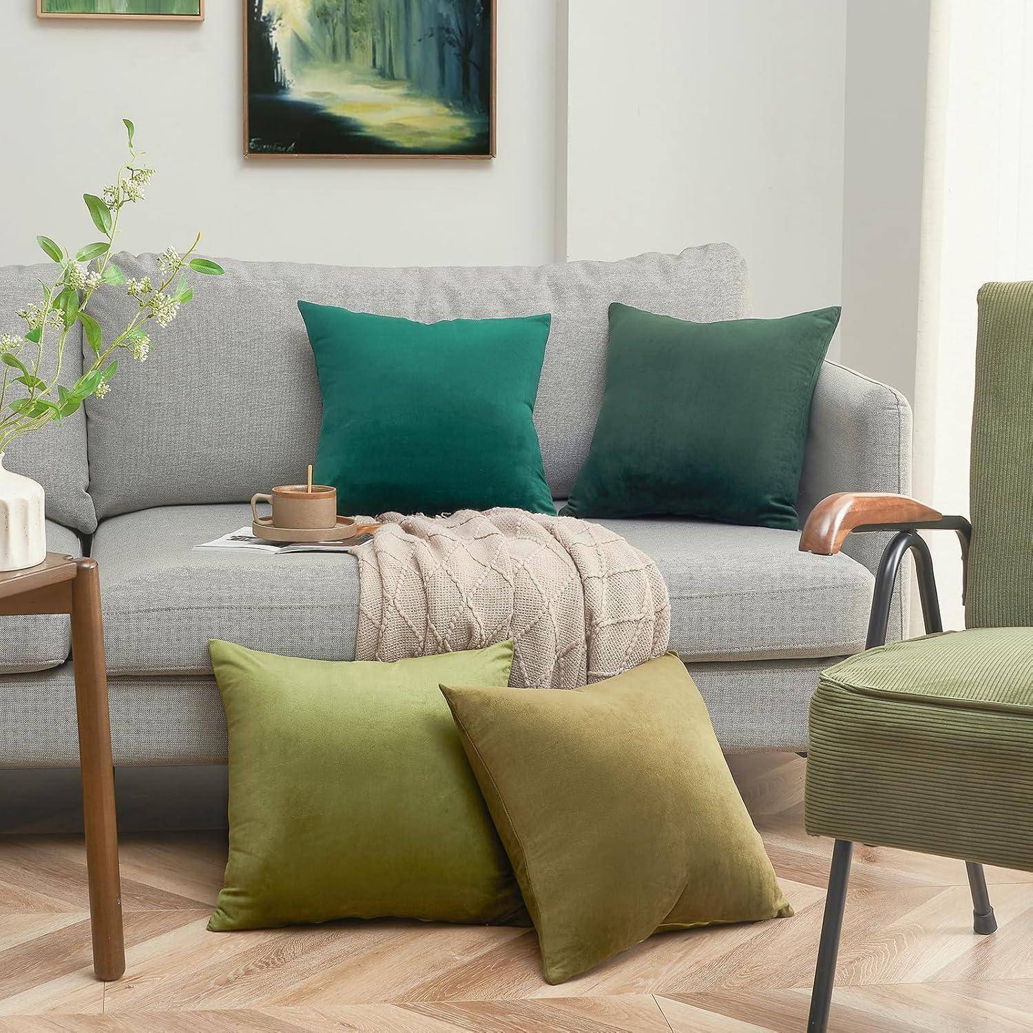 Ein gemütliches Wohnzimmer mit einem grauen Sofa, grünen und türkisfarbenen Kissen, einer beigen Decke, einem Holzseitentisch mit einer Vase und einem serenaden Waldgemälde.