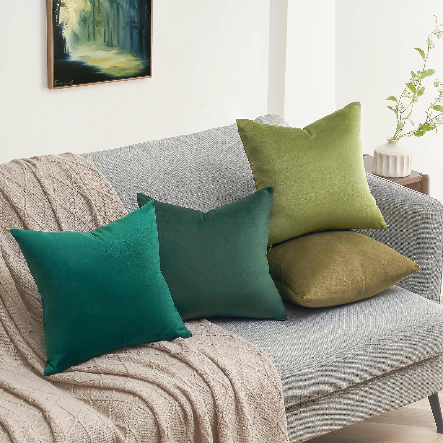 Ein gemütliches Wohnzimmer mit einem hellgrauen Sofa, das mit drei grünen Zierkissen und einer beigen Decke geschmückt ist. Ein kleines Gemälde hängt in der Nähe an der Wand.