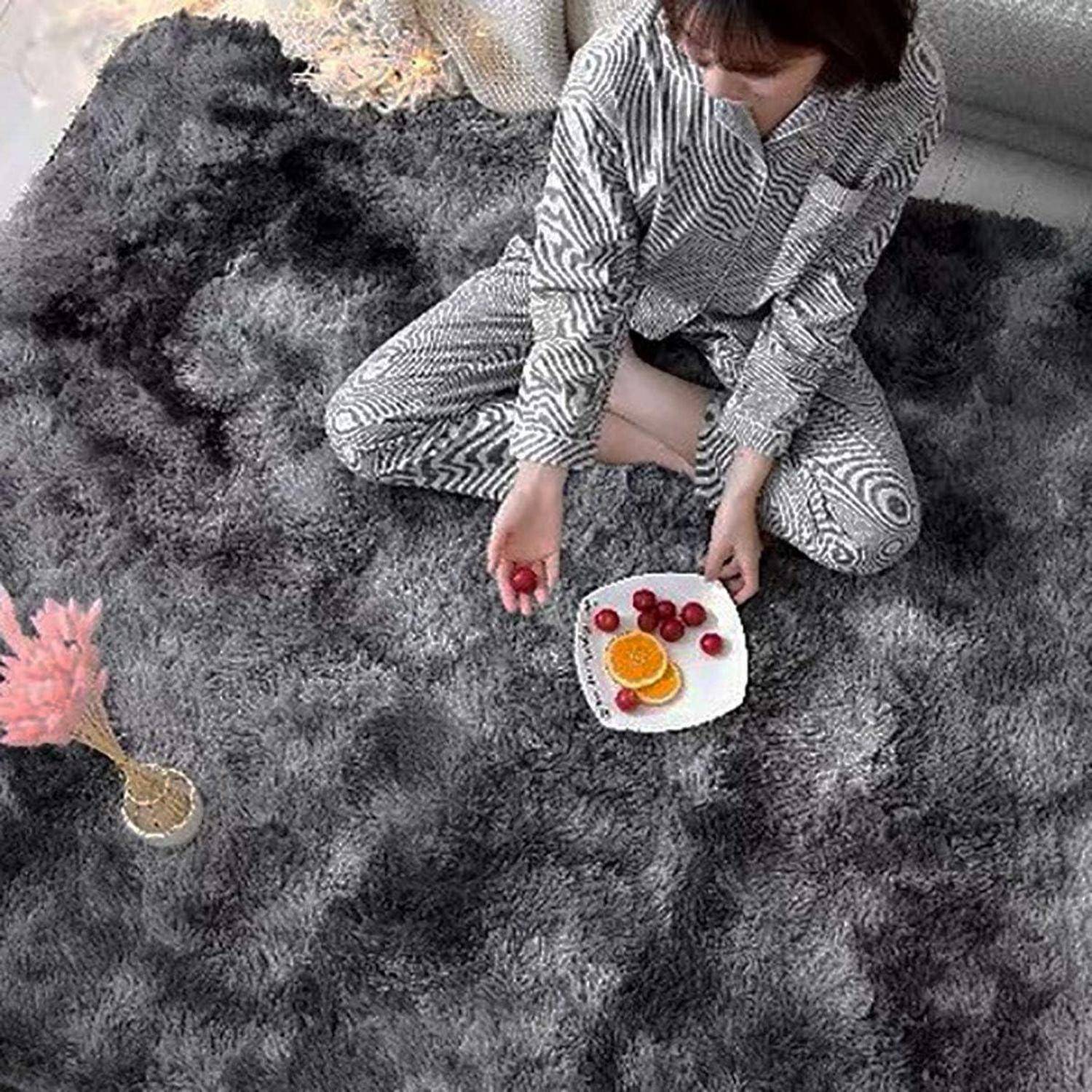 Eine Person in Pyjamas sitzt auf einem grauen, zottligen Teppich und hält einen Teller mit Orangenscheiben, Erdbeeren und einem rosa Getränk davor.
