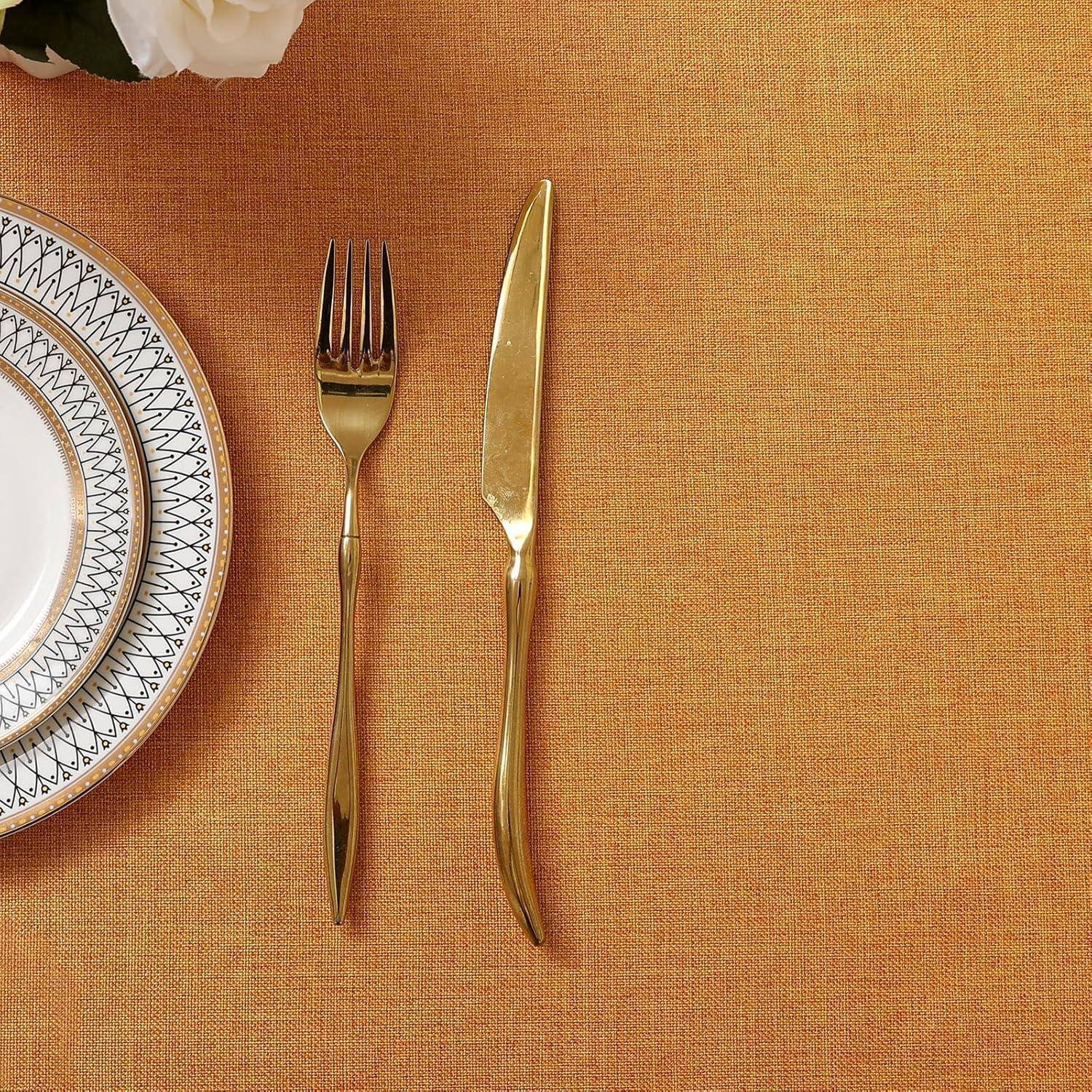 Elegante Tischgedeck mit einem weißen Teller mit gemustertem Rand, einer Gabel und einem Messer auf einem orangefarbenen Tischdecken. Weiße Blumen sind in der Nähe platziert.