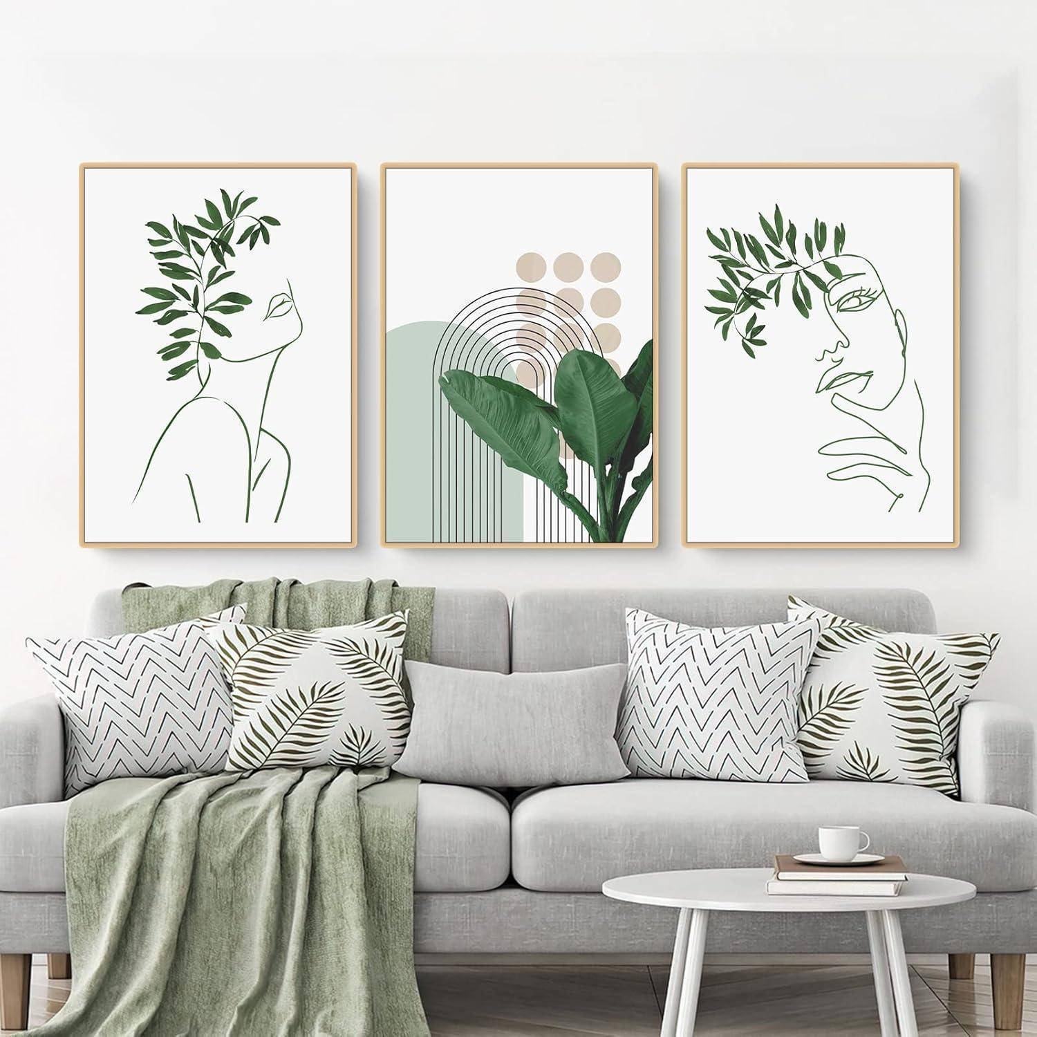 Drei minimalistische Kunstdrucke über einem grauen Sofa zeigen abstrakte Gesichter mit Blattmotiven und geometrischen Formen. Grün gestaltete Kissen und Decke.