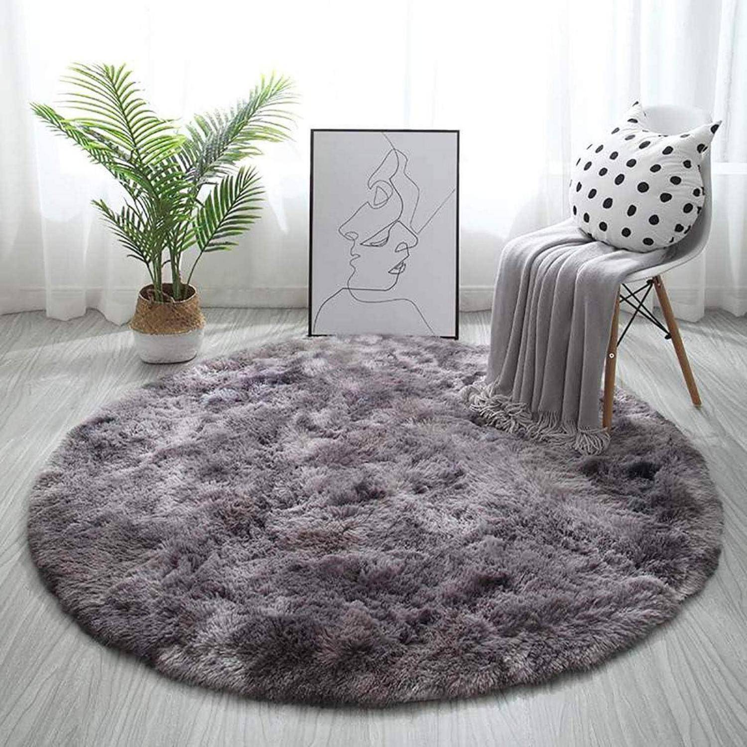 Ein runder, flauschiger grauer Teppich auf einem hellen Holzboden, in der Nähe eines weißen Stuhls mit einer grauen Decke und einem gepunkteten Kissen. Eine Topfpflanze und Linienkunst befinden sich im Hintergrund.