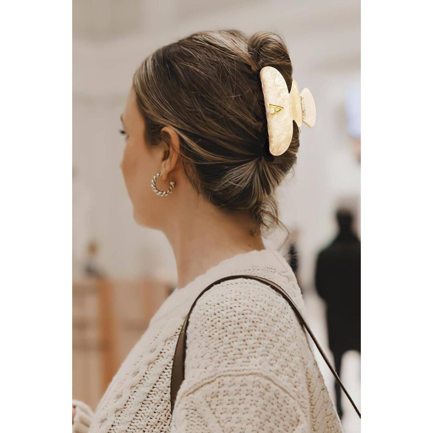 Eine Frau mit hellbraunen Haaren trägt eine große, schmetterlingsförmige Haarspange. Sie ist in einen cremefarbenen Pullover gekleidet und steht in Innenräumen.
