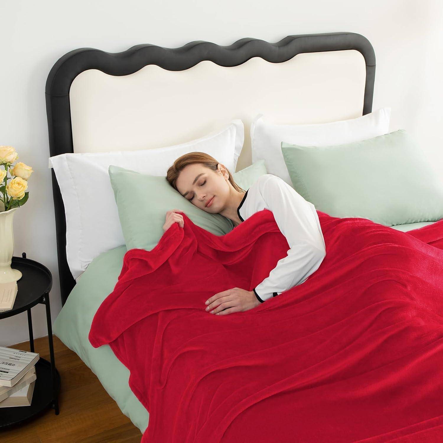 Eine Person, die friedlich im Bett schläft, bedeckt mit einer roten Decke und umgeben von weichen Kissen. Ein kleiner Tisch mit einer Blume steht in der Nähe.